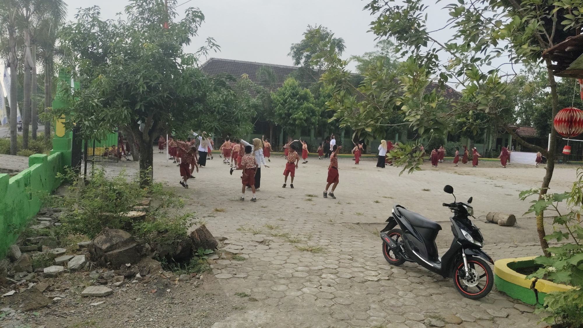 A group of schoolchildren in red and white uniforms are engaged in an outdoor activity, possibly a physical education class or a dance, in the courtyard of a school building in Banjarsari, Indonesia. The courtyard is paved with hexagonal concrete tiles and is surrounded by trees and a low green wall. A black motorcycle is parked in the foreground, to the right of a large tree. The school building, with a tiled roof, is visible in the background, behind the children and trees. The sky is overcast, suggesting a cloudy day.