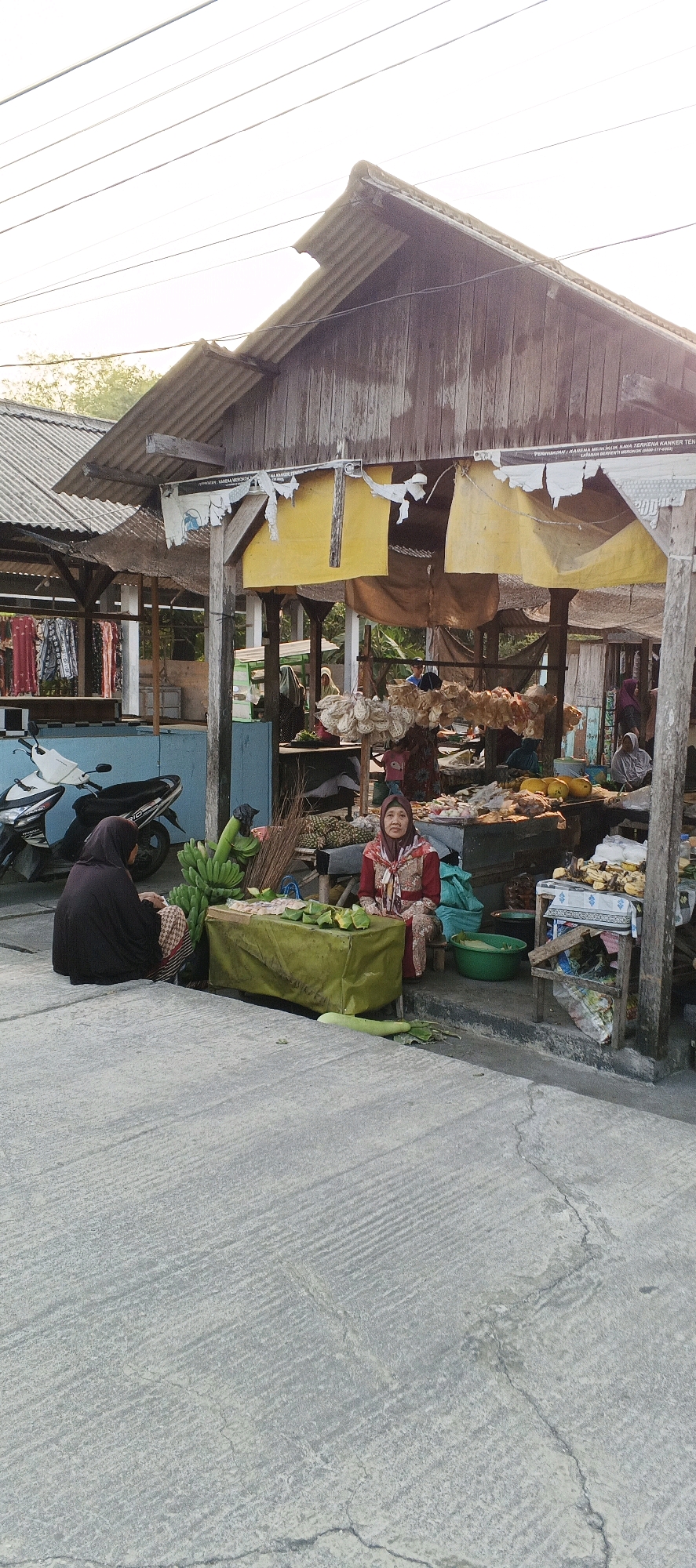 The image depicts an outdoor market scene in Latukan, Indonesia, bathed in the soft light of what appears to be late afternoon or early morning. In the foreground, a gray concrete pathway with a textured, striated pattern stretches across the frame, showing signs of wear and cracking.
To the left, a woman wearing a dark hijab and a patterned skirt is seated on the ground, facing away from the camera, near a bunch of green bananas and a broom. Adjacent to her is a green-covered table laden with what seem to be wrapped food items, possibly snacks or steamed goods, and more of the same green leaves used for wrapping.
To the right of the table, a woman in a red patterned hijab and a matching outfit is seated, facing the camera. She appears to be a vendor. Behind her, a market stall is visible with wooden structures and a weathered corrugated metal roof. Various goods are displayed on shelves and tables within the stall, including what looks like dried food items, fruits like bananas and possibly papayas, and other produce. A green plastic basin is also visible. In the background, other market stalls and people are present, suggesting a bustling marketplace. A blue and white motorcycle is parked to the far left of the image, near some clothing hanging on a rack. The overall impression is of a typical, informal market setting in Indonesia.