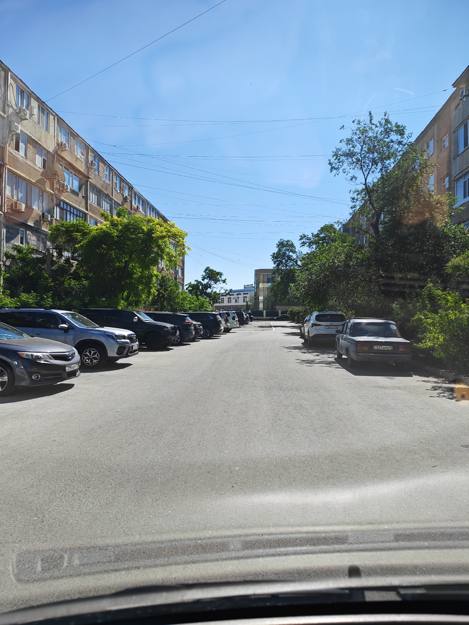 The image depicts a street scene in Aktau, Kazakhstan, on a clear day with a bright blue sky. The viewpoint appears to be from inside a vehicle, looking forward. On the left side of the street are several multi-story apartment buildings with balconies and air conditioning units visible. A row of parked cars lines the street on the left, including a dark gray sedan in the foreground, a silver SUV, and several other vehicles. On the right side of the street, dense green trees and bushes line the sidewalk and the sides of more apartment buildings, some of which appear to be newer construction with more modern architectural styles. Another row of parked cars is visible on the right side of the street, including a light blue sedan with a license plate clearly visible and a white SUV parked behind it. Overhead power lines are strung across the sky, typical of urban environments. The road surface is asphalt. The overall impression is that of a residential street in a city.