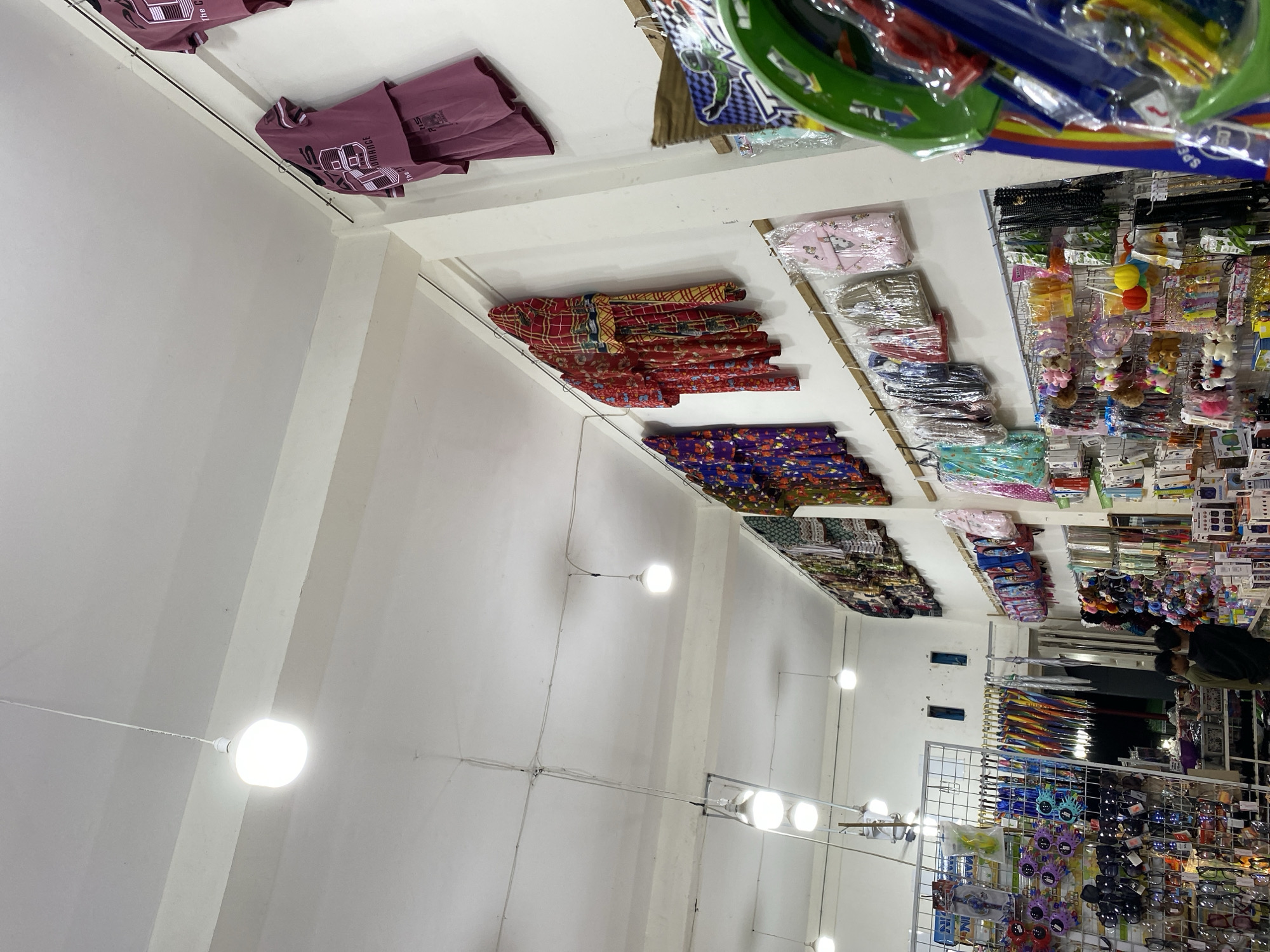 The image shows the interior of a brightly lit store, likely a market or a shop selling various goods. The perspective is from a low angle, looking upwards towards the ceiling and shelves. The scene is densely packed with merchandise. On shelves along the walls, folded textiles, possibly clothing or fabric, are neatly stacked. These items display a variety of patterns, including plaid and floral designs, in vibrant colors like red, yellow, purple, and green.
Above these, on what appears to be a higher shelf, are more packaged items, possibly accessories or toys, with some featuring bright colors and cartoonish designs. In the foreground and extending into the background, metal racks are filled with a wide assortment of products. These include colorful umbrellas, sunglasses with playful designs, small stuffed animals, keychains, and other trinkets.
The store's lighting is provided by several round, white light fixtures attached to the ceiling and walls, creating a well-illuminated environment. In the background, a person wearing a dark jacket is visible, seemingly browsing or working in the store. The overall impression is that of a busy retail space offering a diverse range of affordable items. The context of "Lamteumen Barat, Indonesia" suggests this is a local shop in that region. There is no clear indication of the time of day or weather conditions, as the interior lighting is the dominant light source. Some text is visible on product packaging, but it is too small and blurry to read clearly.