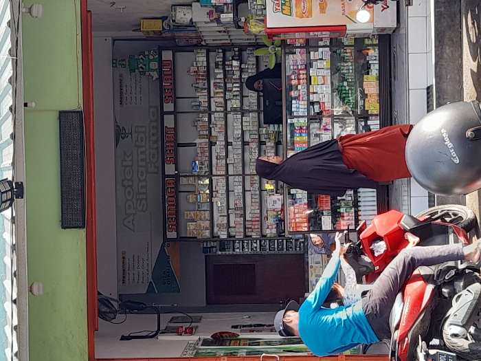 An outdoor scene features a pharmacy, "Apotek Sinambi Farma," with a woman wearing a hijab visible inside. Outside, a man in a blue shirt and cap sits on a red motorcycle, appearing to take a photo with his phone. A helmet rests on the motorcycle seat. The pharmacy's shelves are stocked with medications and other products. The photo is somewhat rotated, requiring the viewer to turn their head slightly to perceive the scene correctly.