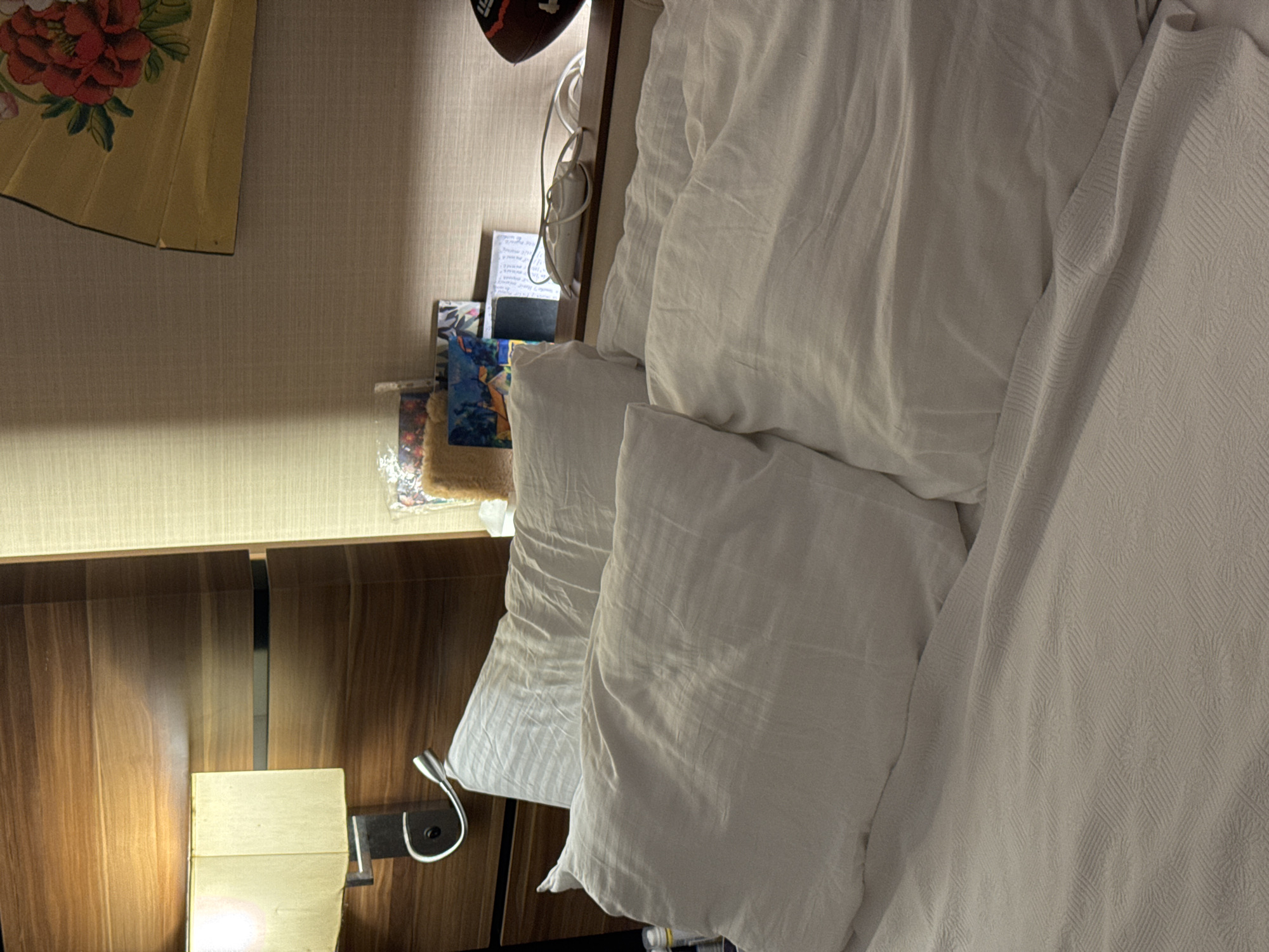 The image shows a close-up view of a made bed with two white pillows, one on top of the other. The pillows are covered in white pillowcases and appear to be made of a soft material. The bed is covered with a white duvet or comforter, and a textured beige blanket is visible in the foreground. To the left of the bed, a bedside lamp with a cream-colored shade is attached to a wooden headboard. The headboard has horizontal wooden panels and a dark horizontal accent. On a shelf behind the pillows, there are several items, including a decorative fan with a floral pattern, a few small framed pictures or cards, and what appears to be a power strip with cords. A football is visible on the right side of the shelf. The background wall is covered with a textured beige wallpaper. The lighting in the room is dim, with light coming from the bedside lamp and an unseen source above. The scene appears to be in a bedroom. The location in Aksu, Turkey is not specified within the image itself.