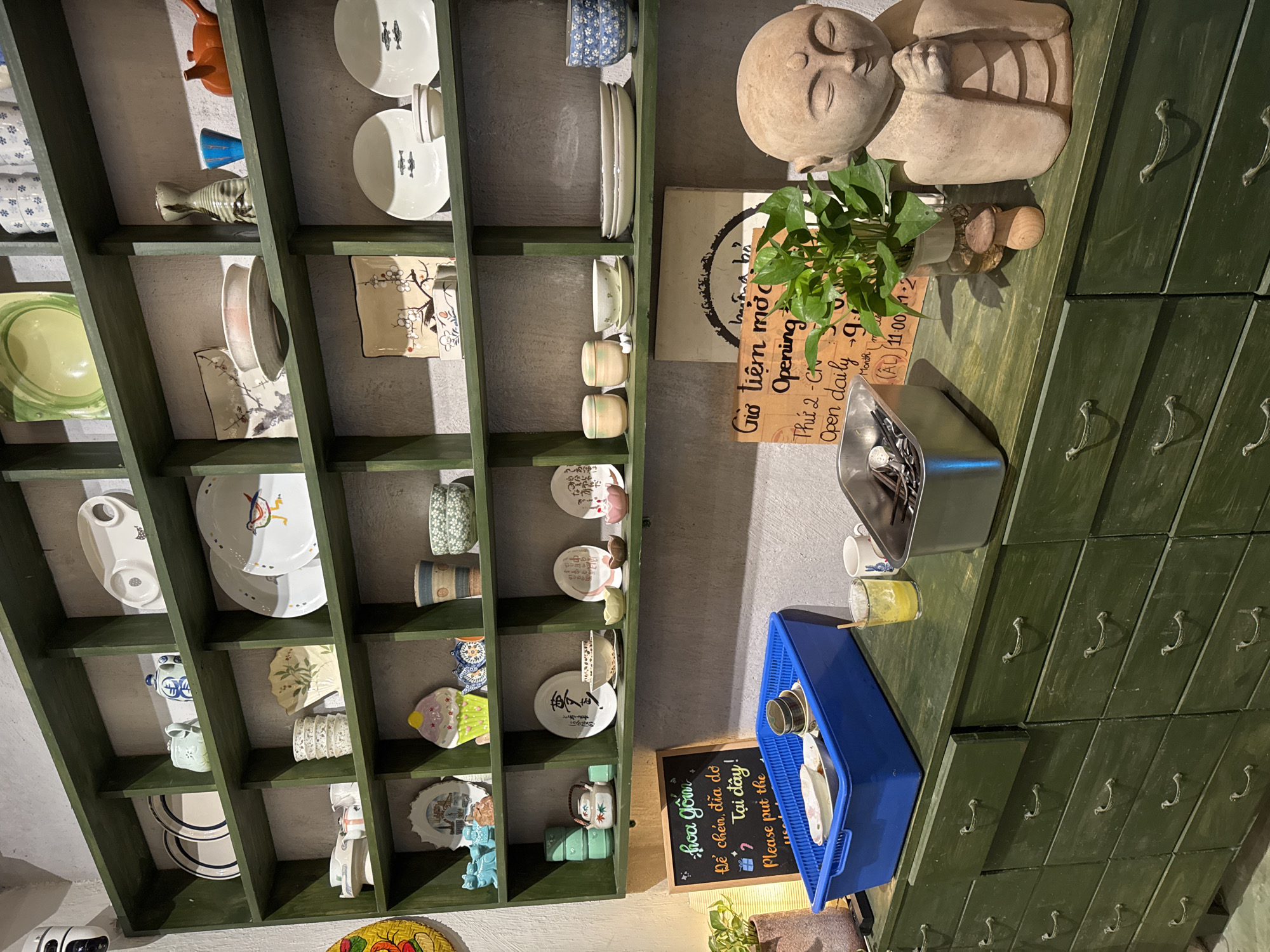 The image shows a vertically oriented display of a shelf filled with ceramics and other items in what appears to be a store or studio, possibly located in Đà Nẵng, Vietnam, given the Vietnamese writing on the chalkboard. The shelves are painted a dark green and contain a variety of plates, bowls, cups, and other decorative ceramic pieces. There is also a statue of a reclining figure, a plant, and a sign with operating hours. Below the shelf, there's a blue plastic container holding dishes and cutlery. A chalkboard contains the text "hoa gốm Để chén, đĩa đơ Tại đây! Please put the" alongside what appear to be decorative elements. The background features a green cabinet with several drawers, suggesting the interior setting of a shop.