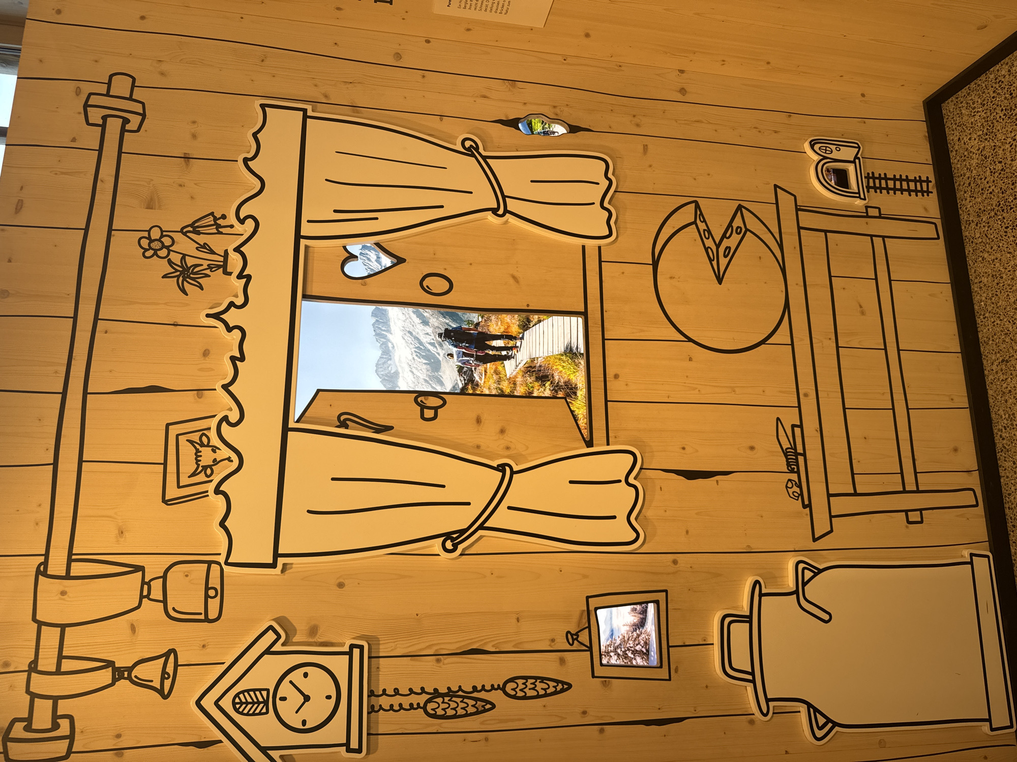 The image depicts a decorative wooden wall in the city of Schattendorf, Switzerland, designed to resemble the interior of a rustic dwelling with cartoon-like illustrations. The wall is adorned with black outlines of various objects, including a cuckoo clock, bells, a milk can, a table with a cheese and knife, and a small mouse door with a miniature railroad track. A prominent feature is a large, stylized window frame with drawn curtains, through which a photograph of two hikers on a wooden boardwalk in a mountainous landscape is visible. The overall scene conveys a sense of alpine charm and a whimsical, storybook aesthetic.