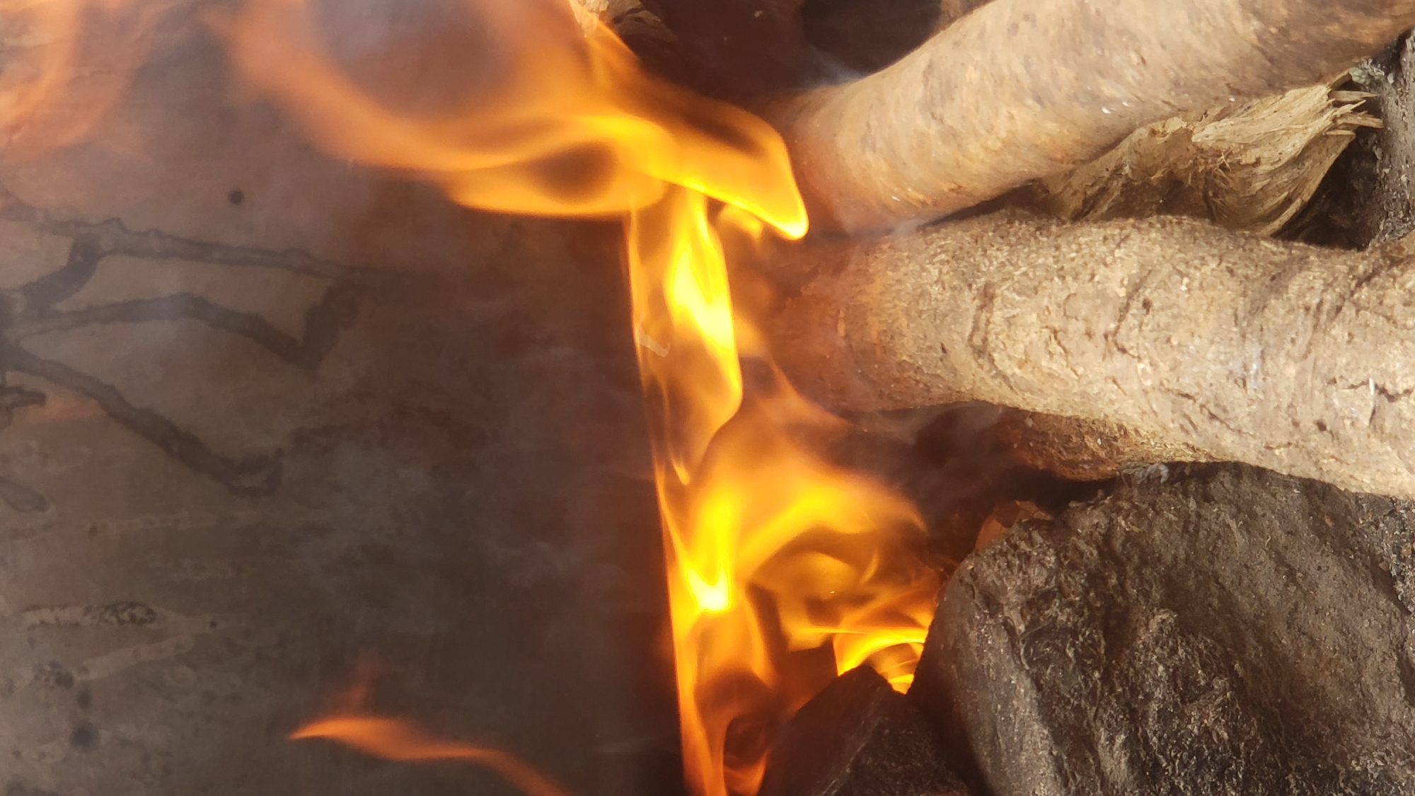 The image depicts a close-up view of a fire burning amidst rough, natural elements. In the foreground, two pieces of firewood, appearing to be substantial logs, are positioned horizontally. To the left, a large, dark-colored rock is visible, partially obscured by the firewood. The fire itself is a vibrant display of orange and yellow flames, with wisps of smoke curling upwards. The background shows a rough, textured surface, possibly a stone wall or an uneven ground, with dark, indistinct markings that could be soot or natural patterns. The overall impression is one of a rustic and uncontrolled fire. The location is identified as Sabon Tasha, Nigeria.