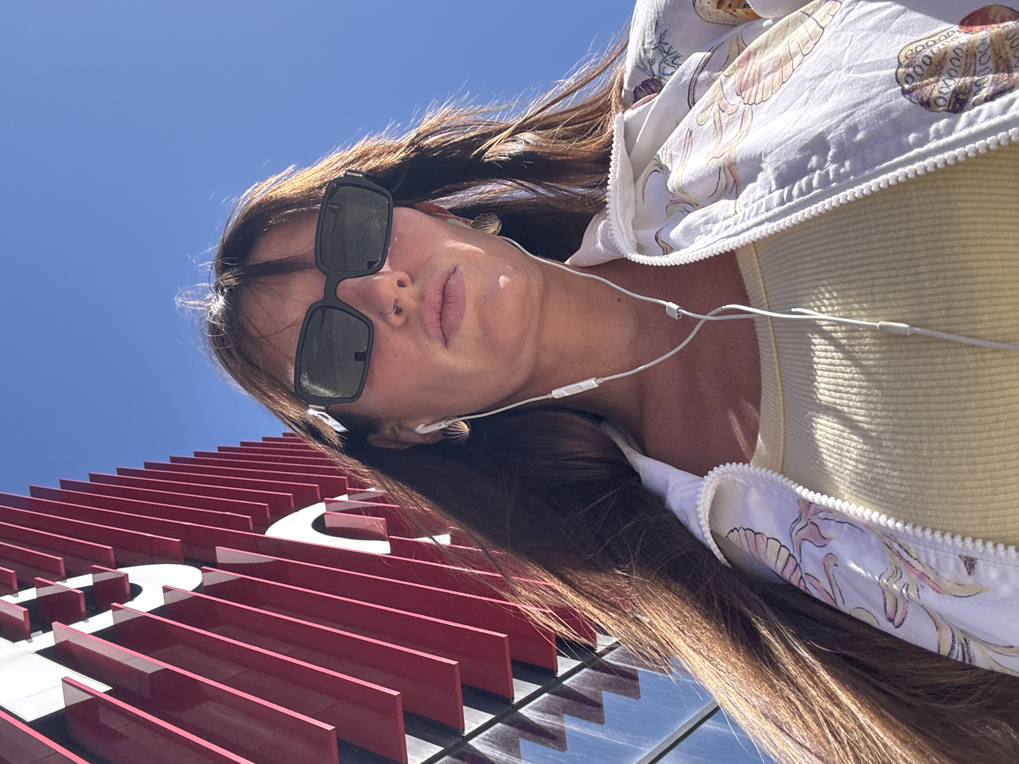 In a low-angle outdoor shot, a woman with long brown hair, wearing large black sunglasses, a light-colored tank top, and a white jacket with a colorful print, stands in front of a modern building with a facade of red and white vertical panels under a clear blue sky. She has white earbuds in her ears and a nose ring. The architecture suggests a contemporary urban setting. While the specific location within Moscow is not identifiable from the visual cues alone, the style of the building is consistent with many modern structures found in the city's commercial and entertainment districts. The lighting indicates a bright, sunny day.