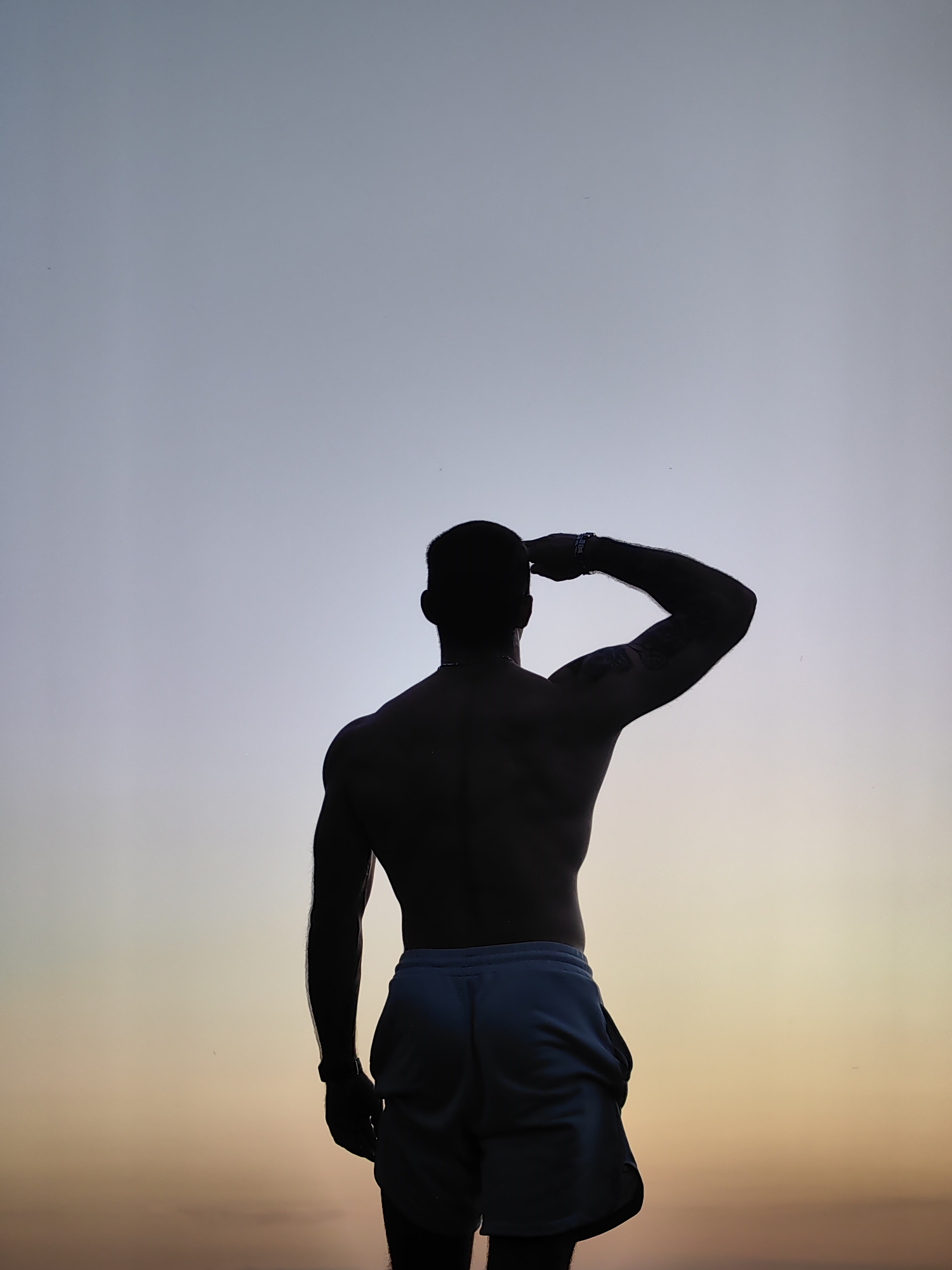The image is a silhouette of a muscular man standing with his back to the camera, looking out at a pale sky. He is wearing light blue shorts. His right arm is raised, with his hand touching his forehead as if saluting or shielding his eyes. He has a tattoo on his right arm. The sky transitions from a pale blue at the top to a soft orange and yellow near the horizon, suggesting either sunrise or sunset. The man is positioned in the lower half of the frame, with the vast expanse of the sky occupying the majority of the image. There are no visible landmarks or specific environmental features that would place this image in Varna, Bulgaria.
