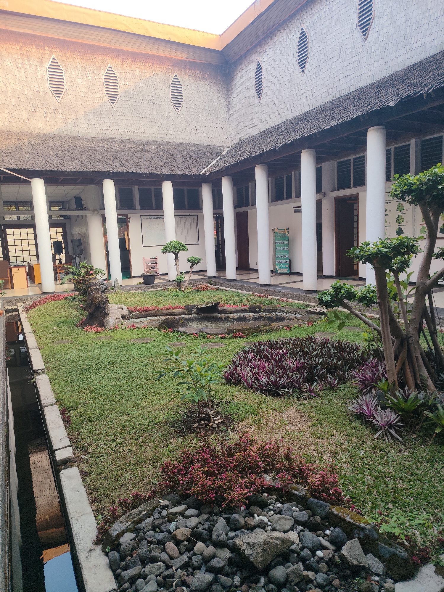 The image depicts an inner courtyard of a building, likely in Tasikmalaya, Indonesia, characterized by traditional architectural elements. A central garden area features lush green grass, ornamental plants with deep purple and pink foliage, and a small water feature with rocks. Several stone pathways wind through the garden. The courtyard is surrounded by a covered walkway supported by large white cylindrical columns. Behind the walkway, the building's facade is visible with wooden framed windows and doors. The roof, made of dark wooden shingles, extends over the walkway and is supported by wooden beams. Several diamond-shaped vents are present on the upper part of the building's exterior walls, beneath the eaves. The lighting suggests it is either early morning or late afternoon, with soft shadows cast across the scene. The overall impression is one of serene, well-maintained outdoor space integrated with a building exhibiting regional architectural influences.