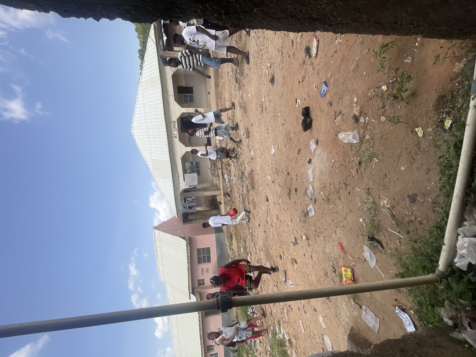 The image depicts an outdoor scene in Lokoja, Nigeria. Several people are visible, some appearing to be walking or standing in a dirt area. The ground is littered with debris, including plastic bags and wrappers. There are buildings present, some with visible signage reading "AS LODGE" and "DECENT LODGE." The sky is blue with some clouds. A metal pipe runs through the lower portion of the frame, partially obscuring the view. The scene appears to be taken from an elevated perspective looking down onto the open space.