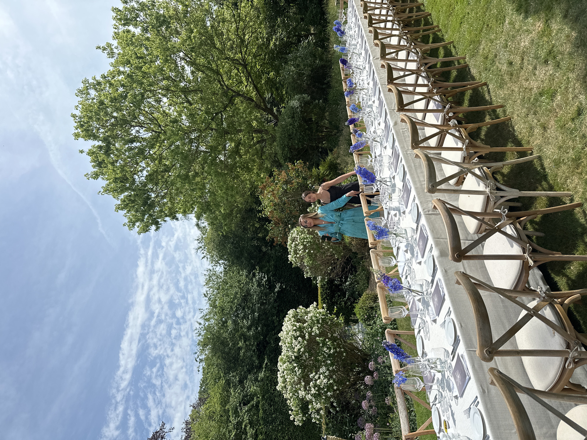 The image shows an outdoor scene with a long banquet table set for a meal. The table is covered with a white tablecloth and has place settings consisting of plates, silverware, glasses, and menus. Blue flowers are placed in vases down the center of the table. There are wooden chairs with off-white cushions set on both sides of the table. Two women are standing near the table, appearing to be interacting or posing for a photo. The background includes green trees, shrubs, and grass, suggesting a garden or park setting. The scene evokes a sense of an outdoor event or celebration. The location is near Cambridge, United Kingdom.