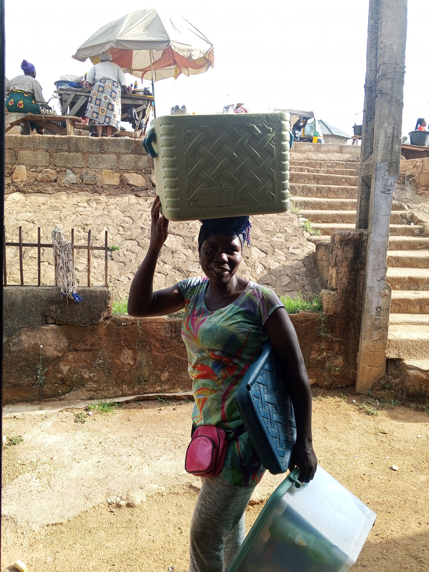 A woman stands on a dirt road in what appears to be an urban setting, potentially in Unknown, Unknown. She is carrying a large green cooler on her head and a blue plastic item in one hand, with a pink bag slung over her shoulder. She wears a colorful patterned shirt and grey pants and appears to be smiling.
Behind her are various structures, including a low stone wall, a metal fence, and steps leading to an elevated area where people and an umbrella are visible. A concrete post with graffiti on it is located to her right, and the surroundings generally suggest a semi-urban or developing area.