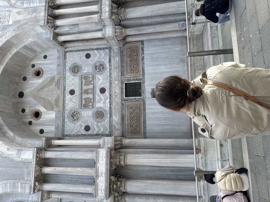 The image displays a detailed view of the St. Mark's Basilica facade in St. Mark's Square, Venice, Italy. In the immediate foreground, a person with dark hair tied in a bun, wearing a beige trench coat and a brown shoulder strap, is viewed from behind, holding a small dark camera or mobile device upwards, presumably photographing the building.
The majority of the frame is occupied by the ornate marble facade of the basilica, characterized by grey and white marble, fluted columns with Corinthian capitals, and arched structures. The facade features extensive decorative elements, including intricate marble inlay patterns, circular dark stone inlays, and rectangular panels with relief carvings. Two prominent round openings are visible within an upper arch, adjacent to a triangular pediment-like design.
Below the main structure, several individuals are present. To the left of the photographer, two people are seated on a stone ledge, one wearing a light-colored puffer vest and the other partially visible. To the right, another individual is squatting or sitting on a lower step, dressed in a dark hoodie and backpack. A metal railing is positioned between the photographer and the seated individuals. The ground is paved with grey stone tiles.