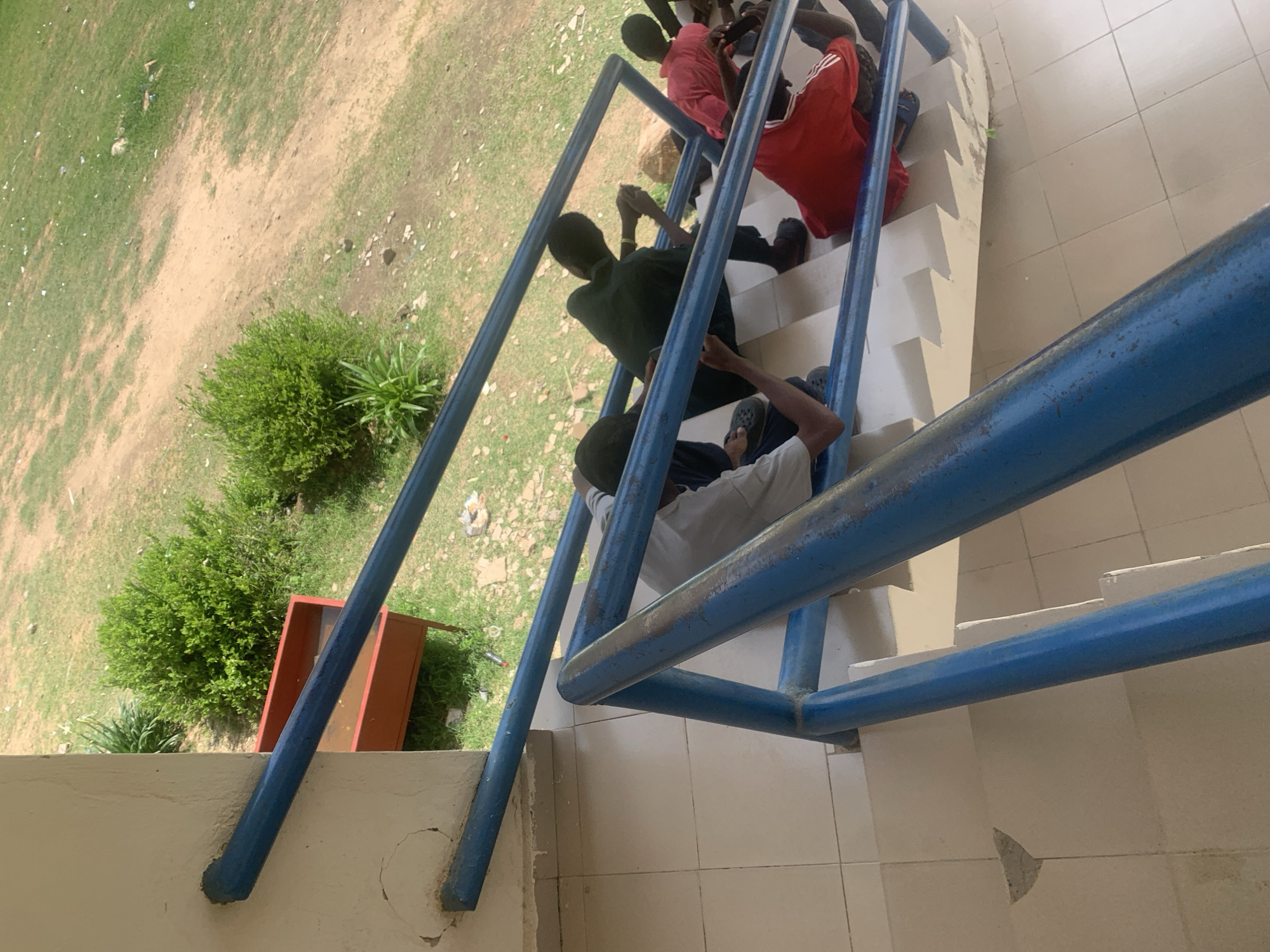 The media file shows a group of young people on an outdoor staircase. The subjects are predominantly male and appear to be teenagers or young adults. They are dressed in casual clothing, with some wearing t-shirts and shorts. One individual is wearing a red shirt with white stripes, and another is in a dark green t-shirt. The staircase has blue metal railings and off-white steps with a light-colored tiled landing.
The individuals are positioned on different steps of the staircase, with some sitting and others standing or leaning. Their postures suggest they are resting or casually interacting. The activity appears to be a moment of downtime or a gathering. One person is looking at their phone, while others seem to be conversing or observing their surroundings.
The setting is outdoors, with a grassy area and a dirt path visible to the left of the staircase. A small, green bush is prominent in the foreground to the left, and a reddish-orange metal container is also present in that area. The ground appears dry and somewhat uneven, suggesting a possibly arid or unmanicured environment. The lighting indicates it is daytime, likely sunny, casting some shadows.
There are no discernible emotions conveyed explicitly, but the casual postures suggest a relaxed atmosphere. The location context of Jalingo, Nigeria, provides a cultural and geographical reference point. No visible text is present in the image.
