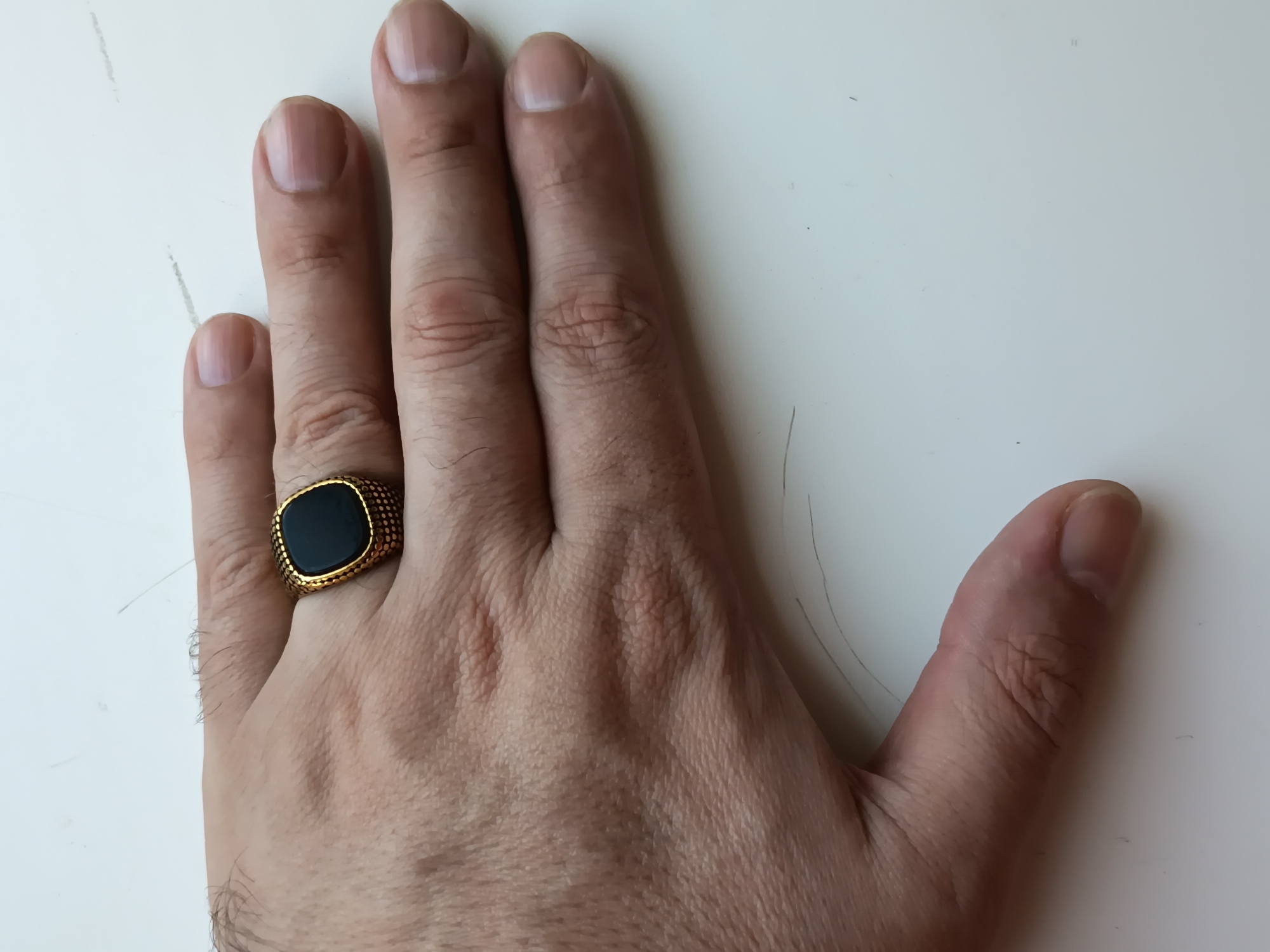 The image shows a close-up view of a person's left hand, positioned on a white surface. The hand is resting with the palm down and fingers slightly spread. A man's gold ring with a black, square-shaped stone is worn on the ring finger. The surface appears to be a table or desk, with faint scratches and a few stray hairs visible. The lighting suggests the photo was taken indoors, possibly in a home or office environment. The image does not contain any information that would allow for the determination of its location within Izmail, Ukraine, or any other specific geographical area.