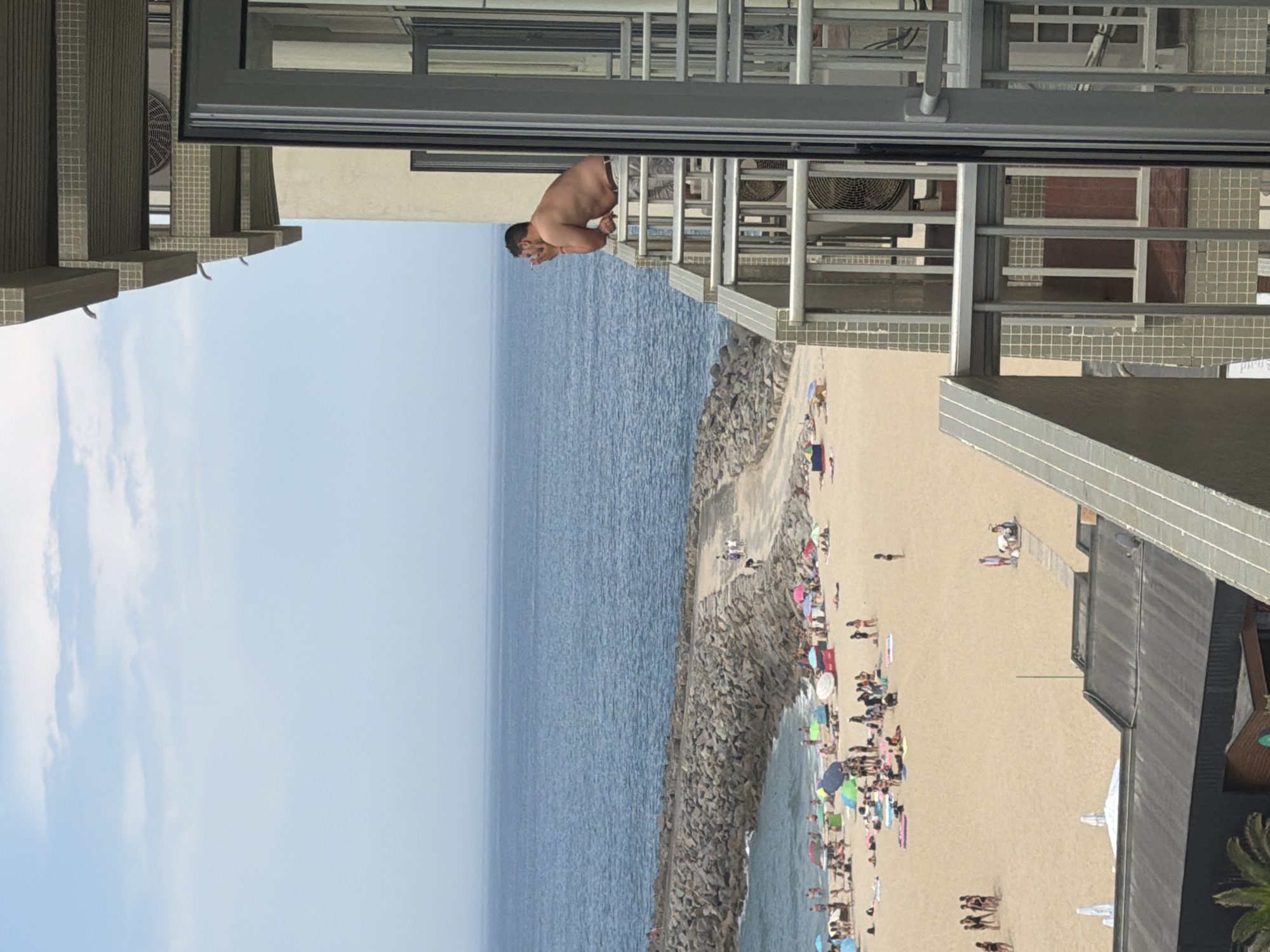 A man is leaning over a balcony railing, looking out at a beach scene. The image is taken from a high vantage point, looking down at the beach and the ocean. The man is shirtless and wearing swim trunks. He is holding something to his mouth, possibly a cigarette or a vape.
The beach is crowded with people, many of whom are sitting on towels or under umbrellas. There are also several boats anchored in the water. The ocean is calm and blue. The weather appears to be sunny and warm. The location is Espinho, Portugal. The image seems to be taken in the late afternoon or early evening, as the sun is starting to set.
The overall mood of the image is relaxed and peaceful. The man on the balcony appears to be enjoying the view, and the people on the beach are enjoying the sun and the sea. The image is a snapshot of a perfect summer day at the beach.