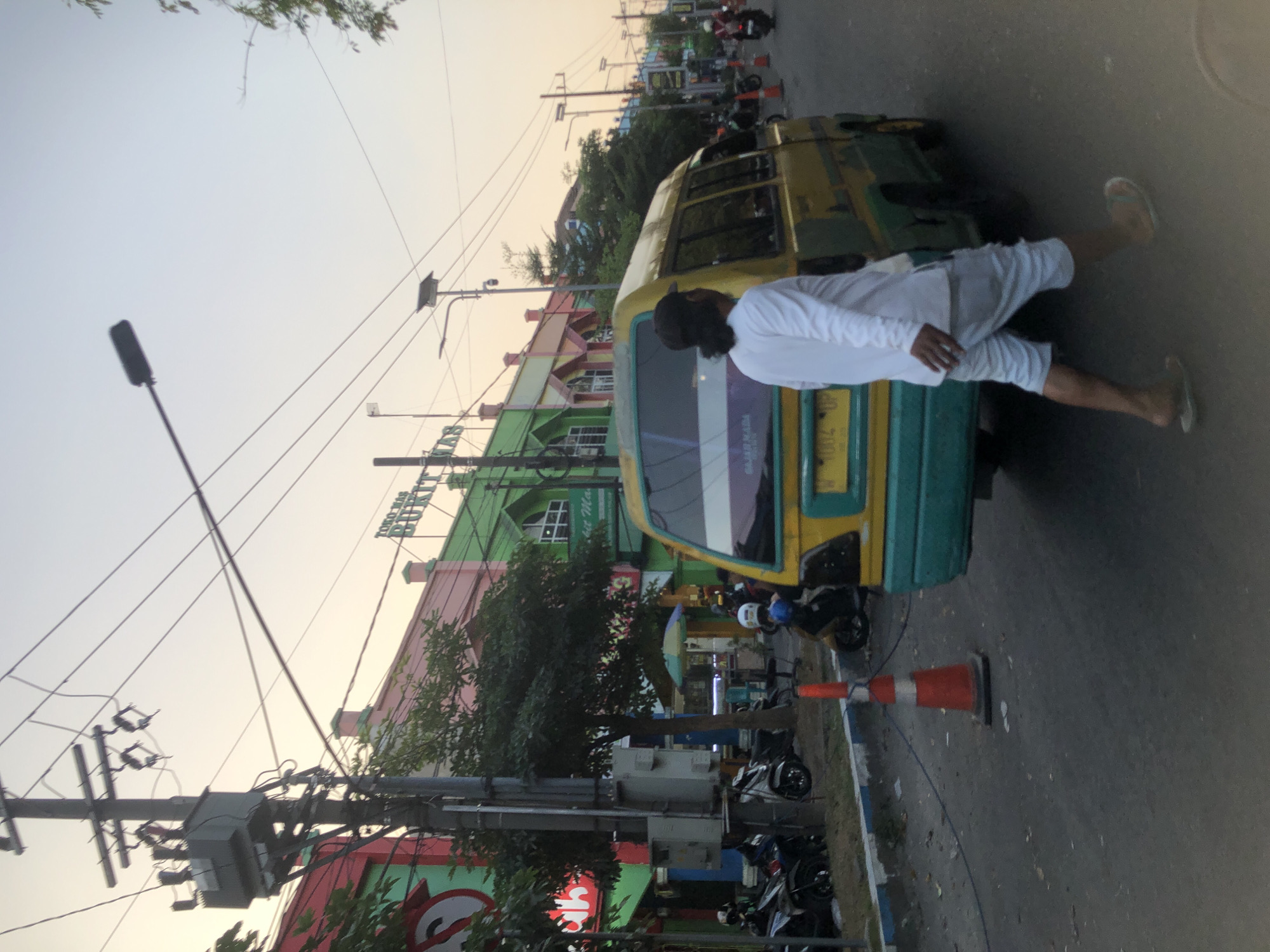 The image depicts a street scene in Larangan, Indonesia, likely during the late afternoon or early evening due to the warm, soft light. In the foreground, a person is walking away from the camera, wearing a white long-sleeved shirt and light-colored shorts, with sandals on their feet. To the left of this individual is a yellow and teal van. A traffic cone, orange with a white band, is positioned on the asphalt road near the van.

The background reveals a row of colorful buildings, predominantly painted in shades of pink, green, and red, with signage that reads "TOKO MAS BUKIT MAS". Trees are visible between the buildings and along the sidewalk. Various motorcycles are parked or moving on the side of the road. Overhead, electrical poles and wires crisscross the sky, which is a pale orange and blue gradient, suggesting either sunrise or sunset. Streetlights are also present. The road surface is asphalt, with a blue and white curb on the left side of the street.