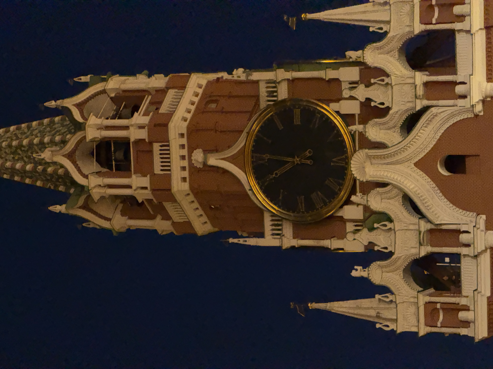 The image shows a close-up of the Spasskaya Tower in Moscow, Russia, at night. The tower is made of red brick and features ornate white decorative elements. A large clock with Roman numerals is visible on the tower. The sky is dark blue. There are no people or animals visible in the frame.