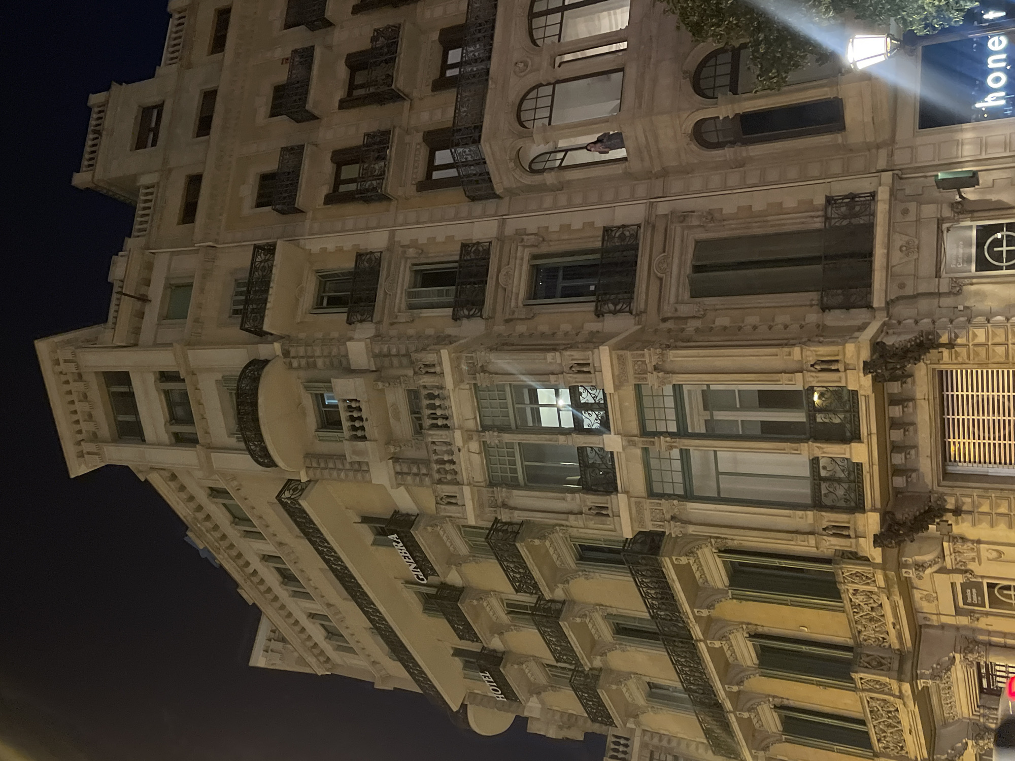 The image shows a nighttime view of the Hotel GIN EBRA in Barcelona, Spain. The hotel is a multi-story building with ornate architectural details, including balconies with wrought-iron railings and decorative stone carvings. The hotel's name is illuminated in large letters on the facade. The building is adjacent to another similar building, creating a dense urban streetscape. A street lamp is visible in the lower right corner, casting a warm glow on the scene. A person can be seen standing on a balcony in the background. The sky is dark, indicating it is nighttime.