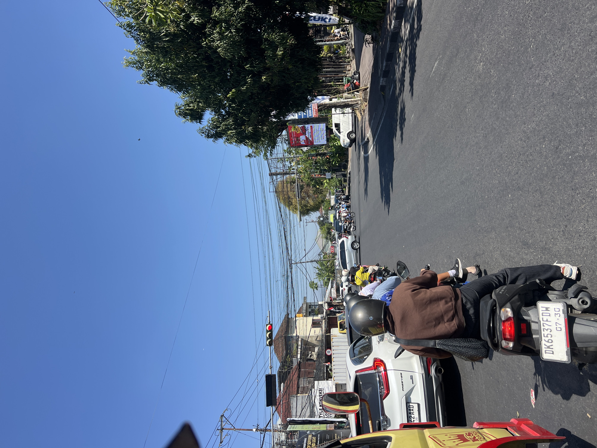 The image shows a busy street scene in Denpasar, Indonesia, on a sunny day with a clear blue sky. The perspective is from behind a motorcycle rider, looking forward. In the foreground, a person wearing a brown jacket and striped pants is riding a black scooter, with a black woven bag slung over their shoulder. Their license plate reads "DK6537FDW 07.30".
Traffic is moderate, with several other motorcycles and a white car in front of the scooter. The white car's license plate is partially visible, reading "42AAD". To the left, a yellow vehicle, possibly a truck or van, is also in traffic. Further down the road, there are more vehicles, including a white van and numerous motorcycles, suggesting a bustling urban environment.
Buildings line the street, some appearing older with weathered facades and tiled roofs. Overhead, a dense network of electrical wires crisscrosses the sky, a common sight in many Indonesian cities. A traffic light is visible, with the red light illuminated, indicating that traffic is stopped.
Vegetation, including trees with lush green foliage, grows along the sidewalk and behind some of the buildings, providing a contrast to the urban infrastructure. Billboards and signs are present on some of the buildings, advertising products or services. The road surface is asphalt, showing signs of wear and tear. The overall impression is of a typical day in a lively Indonesian city, with a constant flow of traffic and a vibrant street life.