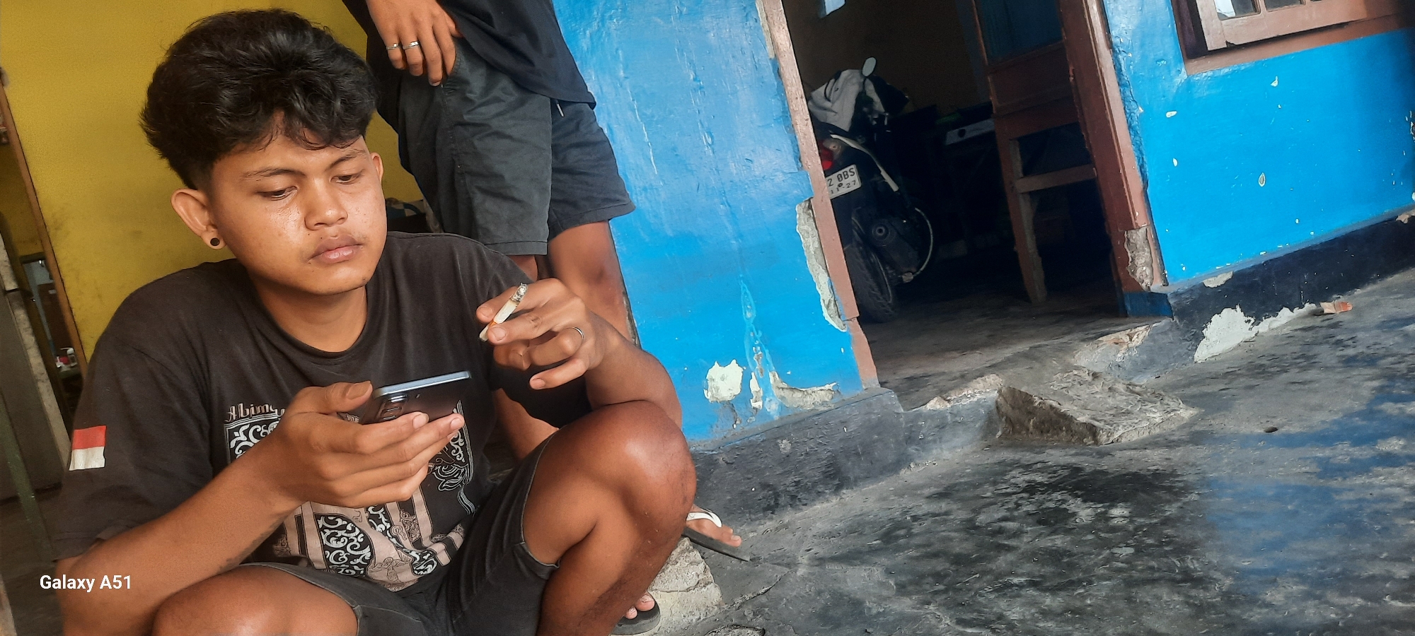In an outdoor scene, a young man, likely of Indonesian ethnicity, is crouching and focused on his mobile phone. He is wearing a black t-shirt with a white and red flag emblem on the left sleeve and a graphic print on the front. He holds a lit cigarette between his fingers, close to his mouth. His right hand holds the mobile phone, and his left hand is raised, holding the cigarette. He has short, dark, curly hair and appears to be wearing black shorts. Another person, partially visible in the background, is standing and wearing black shorts and a t-shirt. They have rings on their fingers.
The environment appears to be a semi-urban or rural area, with a brightly painted blue wall that is peeling in places. The ground is made of concrete, with some debris and cracks. To the right, an open doorway reveals a darkened interior where a motorcycle is parked. The scene evokes a casual, perhaps slightly dilapidated, setting. The presence of the Galaxy A51 logo in the lower left corner indicates that the image was captured by a Samsung Galaxy A51 mobile phone. While the specific location within Selopuro, Indonesia, is not visually identifiable, the overall context suggests a residential or communal outdoor space.