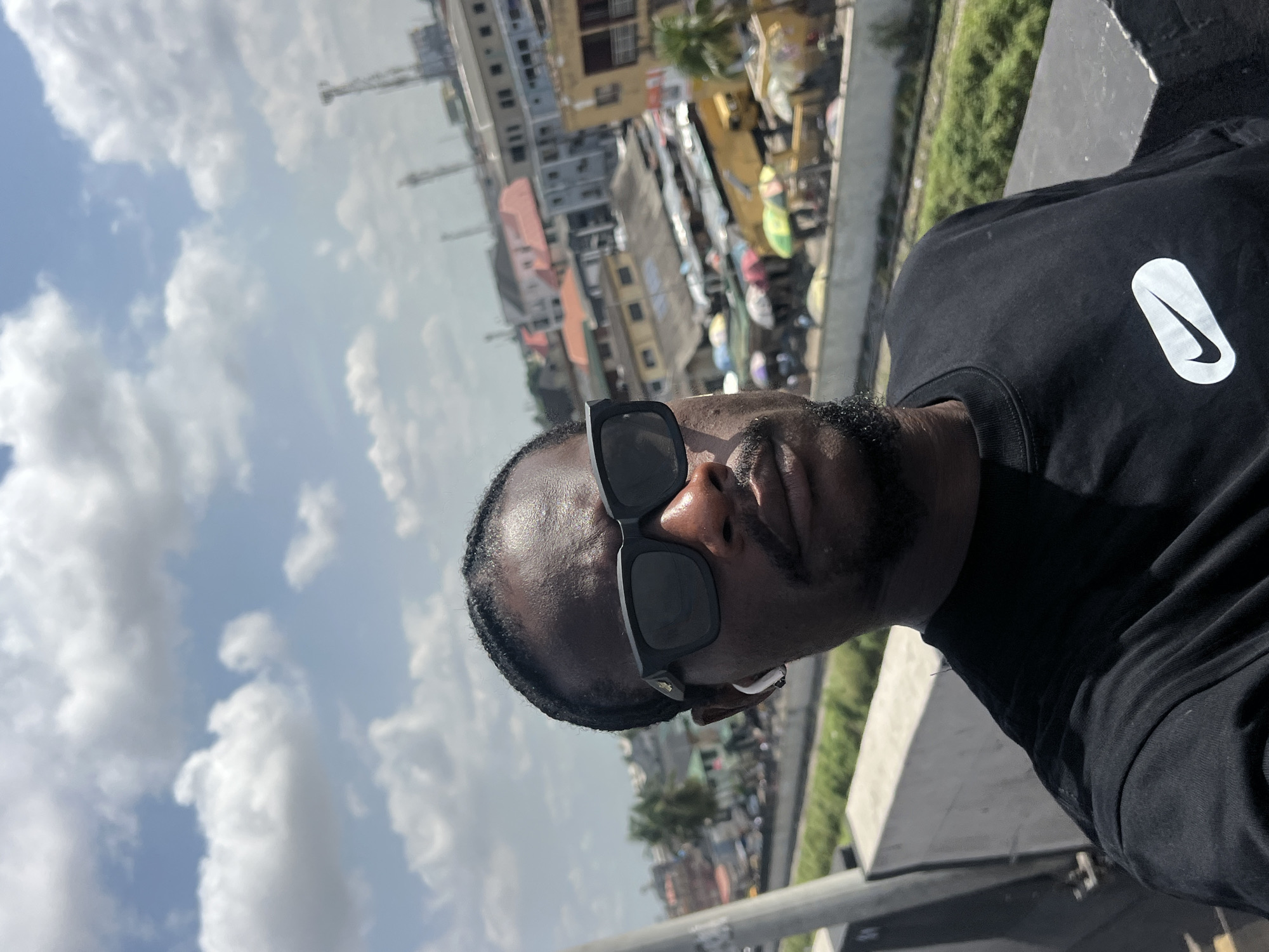 The image is a vertical, selfie-style photograph, rotated 90 degrees clockwise, featuring a man outdoors in an urban environment. The subject is a dark-skinned man, captured from the chest up, looking towards the camera. He wears black, rectangular-framed sunglasses that cover his eyes and part of his nose. A short beard or stubble is visible on his chin, and a white earbud is inserted into his right ear.
He is dressed in a plain black crew-neck t-shirt. On the left side of his chest, a prominent white logo is visible, resembling an elongated oval containing a black Nike swoosh.
The background depicts a vibrant urban landscape, identified as Ikeja, Nigeria. Numerous buildings of varying sizes and architectural styles are densely packed, with some showing weathered facades. A body of water or a canal, partially lined with green vegetation, can be seen winding through the cityscape. The man appears to be positioned on an elevated structure, such as a bridge or overpass, providing a vantage point over the city below. The sky is bright blue with scattered white cumulus clouds, indicating a clear and sunny daytime. The overall impression is a casual portrait taken on a bright day in a bustling city.