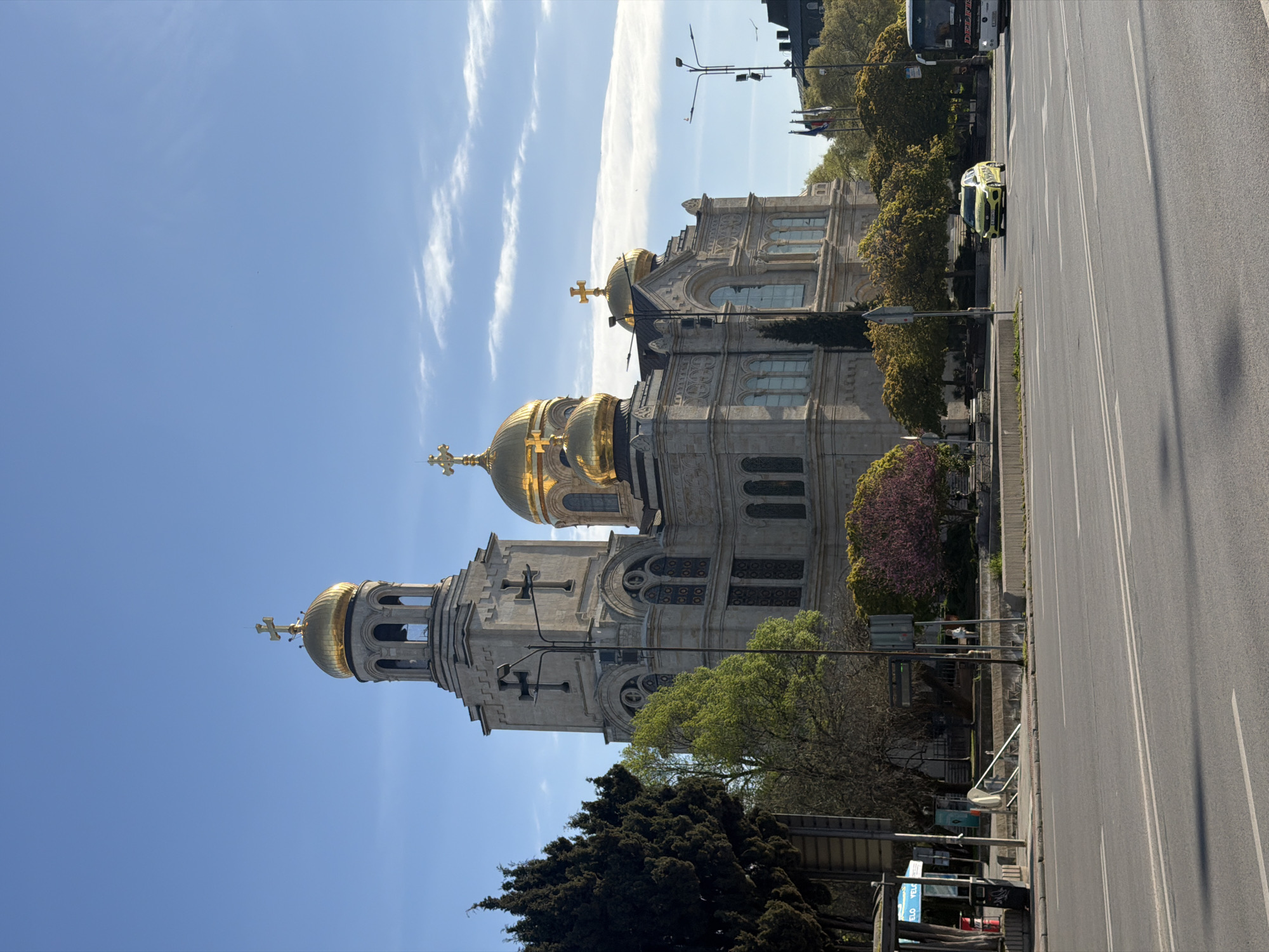 The image shows the Dormition of the Mother of God Cathedral in Varna, Bulgaria, from a street-level perspective. The cathedral's distinct gold domes are prominent against a blue sky with wispy clouds. The architecture is intricate with decorative features, windows, and crosses. The foreground includes a road with painted lane markings, and a bus and car are seen on the right. Trees with spring foliage partially obscure the base of the cathedral. A bus stop is visible in the lower left corner.