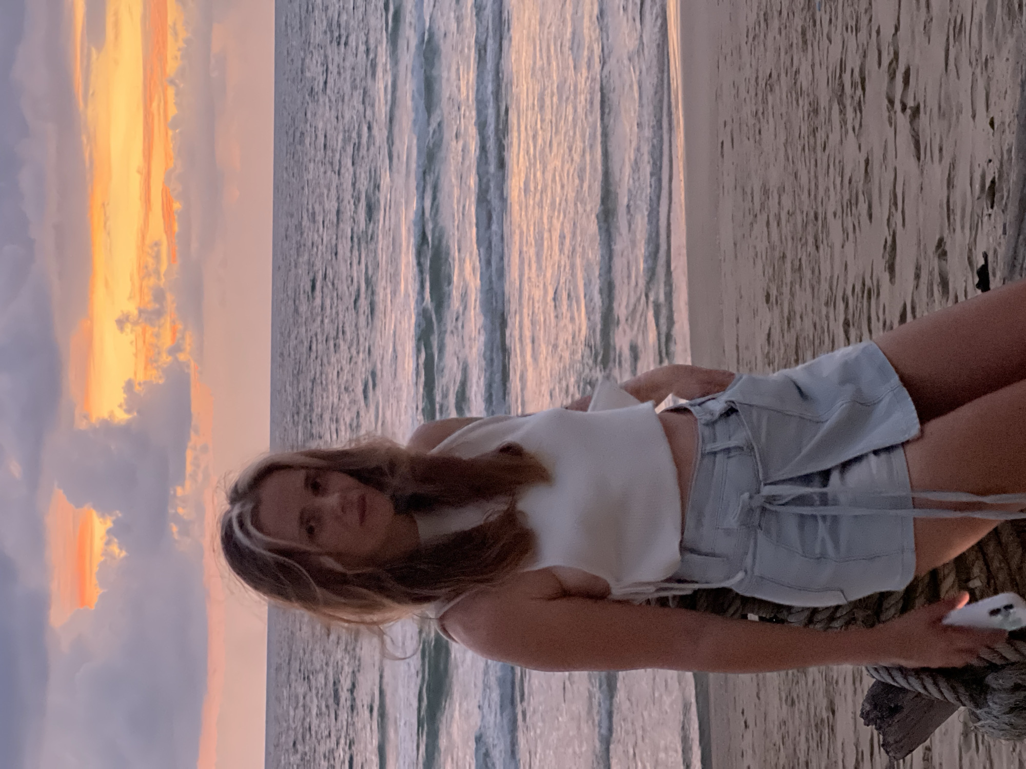 A woman stands on a beach in Choeng Thale, Thailand, with the ocean and a sunset in the background. She is wearing a white cropped top and a light blue wrap-around skirt. Her left hand is in her pocket, and her right hand is holding a white smartphone. The sky is filled with pink, orange, and purple clouds, and the ocean waves are gently breaking on the shore. The sand is a light brown color. There is a thick rope and a wooden post to her left.