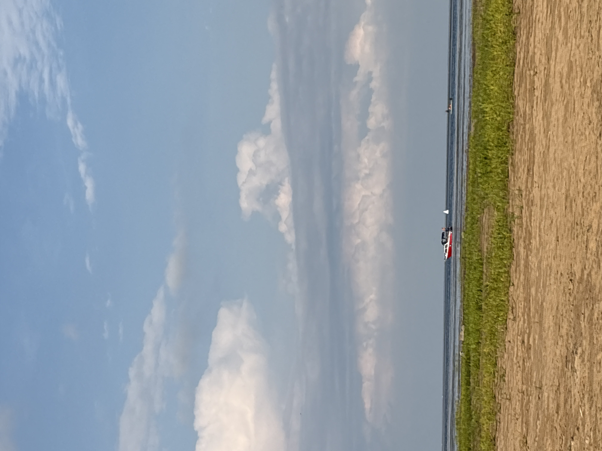 The image depicts a tranquil scene of the Volga River in Koshkino, Russia. The foreground is a sandy beach with green grass growing at the water's edge. The river stretches out to the horizon, with a few small boats visible. In the middle ground, a red and white motorboat is cruising on the water, accompanied by a small white sailboat. Further in the distance, another small vessel can be seen. The sky above is a clear blue, dotted with fluffy white clouds. The overall atmosphere is peaceful and serene, suggesting a pleasant day at the river.