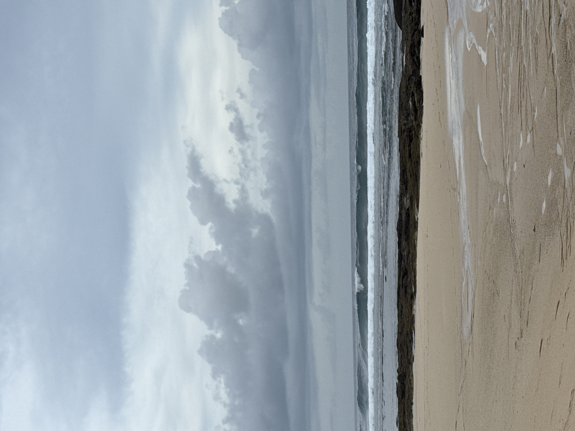 The image depicts a beach scene in Pecatu, Indonesia, under a cloudy, overcast sky. A stretch of wet sand dominates the foreground, marked with patterns likely left by receding waves. To the left of the sand is a dark, rocky area followed by the ocean, with white-capped waves. The sky is filled with heavy gray clouds.