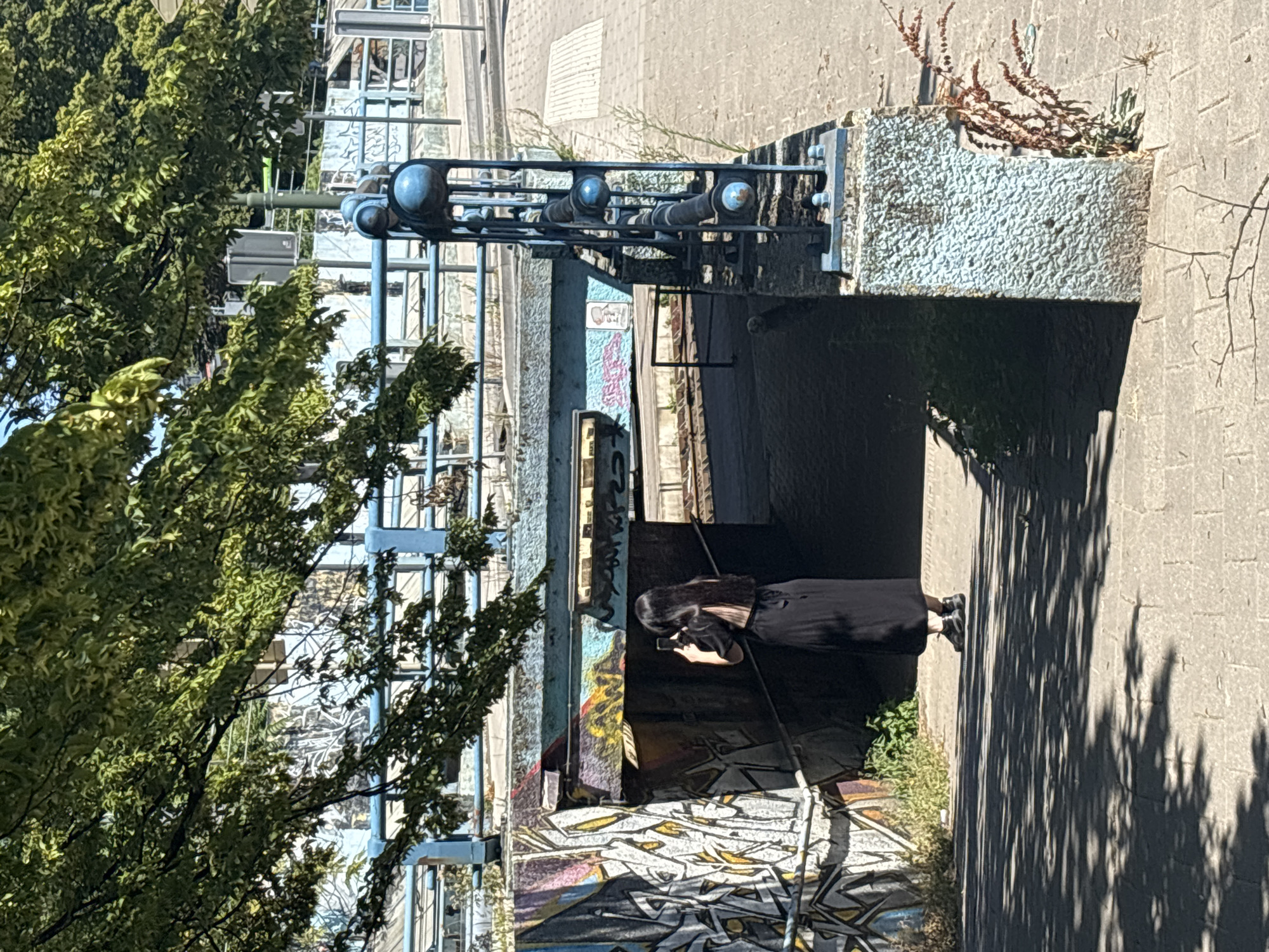 A person with long black hair wearing a black dress stands in a shadowed area with graffiti on the walls. They are holding a phone up, seemingly taking a picture. The area appears to be an underpass or walkway, with a concrete structure overhead adorned with some blue pipes and light fixtures. To the left of the scene, a large tree with green leaves partially obscures a blue metal structure, possibly part of a bridge or walkway. The ground is paved with cobblestones and there are patches of dry grass and plants. Sunlight casts dappled shadows on the ground and wall, suggesting it is daytime. The graffiti on the walls is colorful and abstract. The overall mood is somewhat urban and gritty, with a hint of artistry due to the graffiti. The setting, with the combination of urban infrastructure, graffiti, and natural elements like the tree, suggests a public space in a city. The presence of a phone and the act of taking a picture imply a moment of personal observation or documentation.
