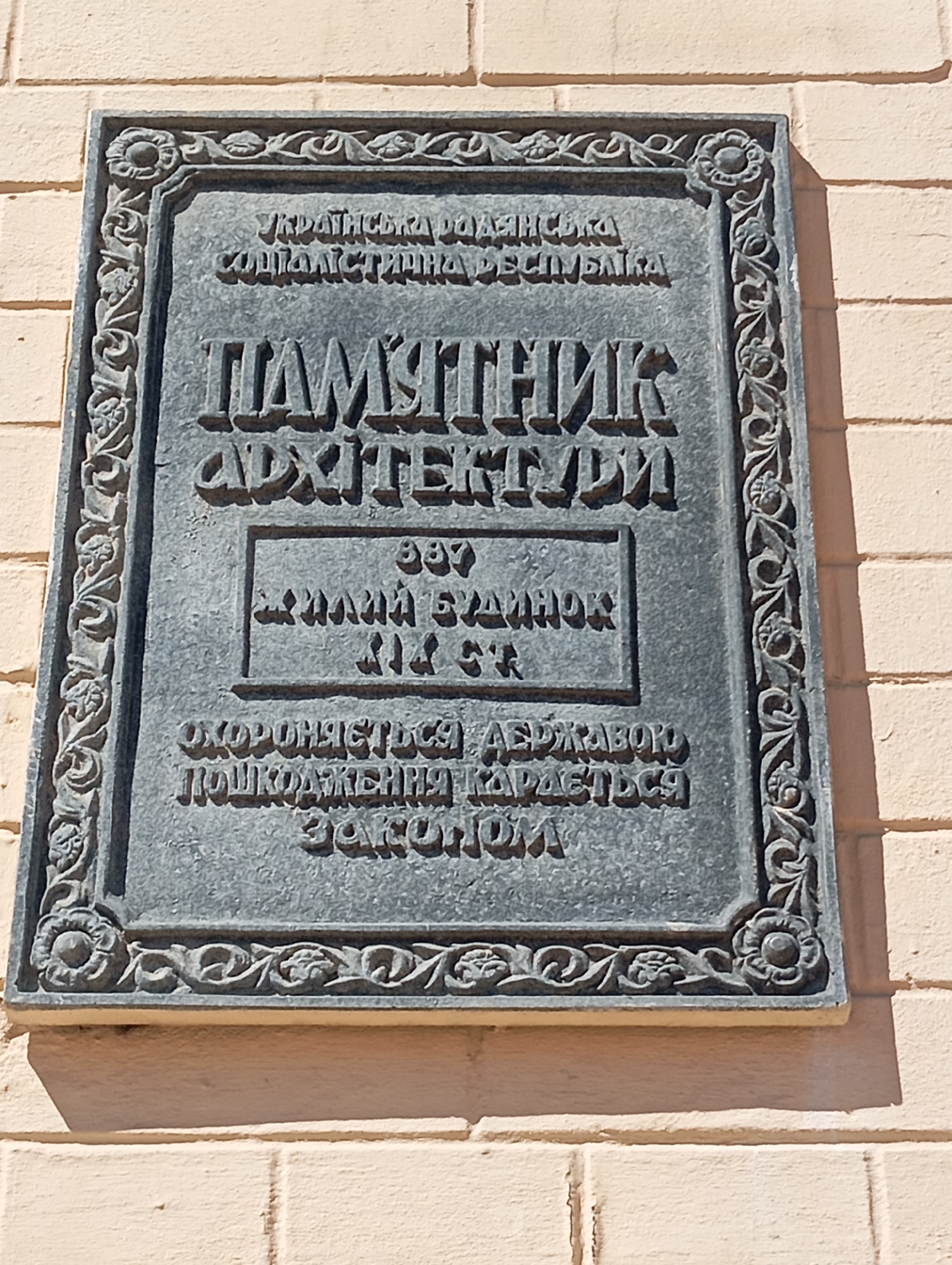 A weathered, dark gray plaque with ornate borders is affixed to a beige brick wall. The plaque, which appears to be made of stone or metal, has raised, embossed lettering. The top line of text reads "УКРАЇНСЬКА РАДЯНСЬКА СОЦІАЛІСТИЧНА РЕСПУБЛІКА" (Ukrainian Soviet Socialist Republic). Below this is a large inscription that reads "ПАМ'ЯТНИК АРХІТЕКТУРИ" (Monument of Architecture). Centered within this is a smaller rectangular section containing the year "1997" and then lines of text that are difficult to fully decipher but appear to be "ЗНАЧНІЇ БУДИНОК" (significant building) and potentially a building number or designation. At the bottom, a final line of text states "ОХОРОНЯЄТЬСЯ ДЕРЖАВОЮ. ПОШКОДЖЕННЯ КАРАЄТЬСЯ ЗАКОНОМ" (Protected by the state. Damage is punishable by law). The plaque is mounted centrally on the wall, casting a slight shadow to the right. The brickwork of the wall is uniform in color and texture. The overall scene suggests an urban environment, likely in Kyiv, Ukraine, given the Ukrainian language on the plaque and the architectural preservation notice.