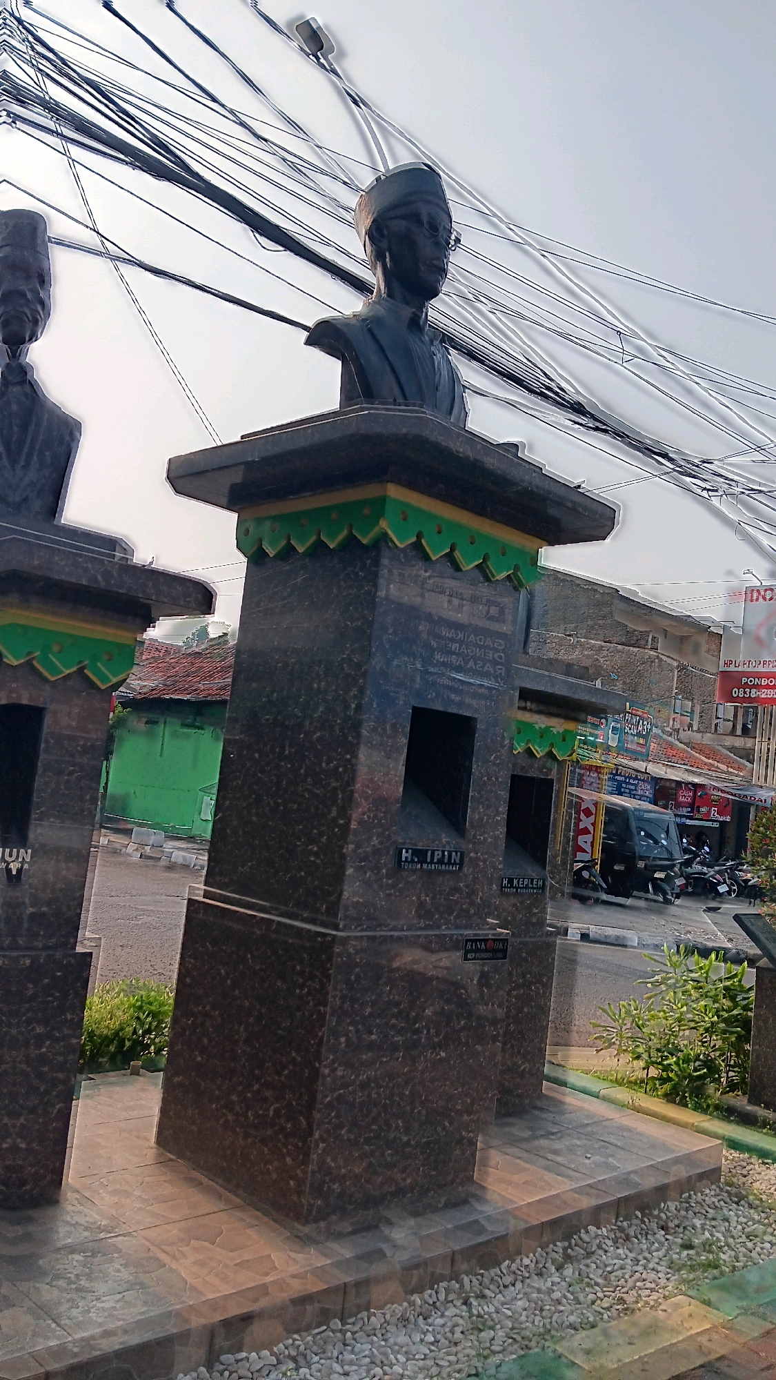 The image shows a bust of a man wearing glasses and a cap, with a prominent inscription on the pedestal below him. The inscription, partially obscured, appears to read "H. IPIN TOKOH MASYARAKAT" which translates to "H. IPIN COMMUNITY FIGURE". The bust is made of dark material, possibly bronze or stone, and stands on a dark granite pedestal decorated with green and yellow trim.
To the left of this bust, another bust is visible, also on a similar pedestal. The inscription on this pedestal is largely unreadable. The busts are positioned outdoors, in what appears to be a public space. In the background, a green building with a red-tiled roof is visible, along with a street with vehicles, including a three-wheeled auto-rickshaw (bajaj) and motorcycles. There are also shops with signs and overhead electrical wires.
Based on contextual information often associated with this image, this scene is located at the "Taman H. Ipin", a park in Cipete Utara, Kebayoran Baru, South Jakarta, Indonesia. The park is dedicated to H. Ipin, a respected community figure in the area.