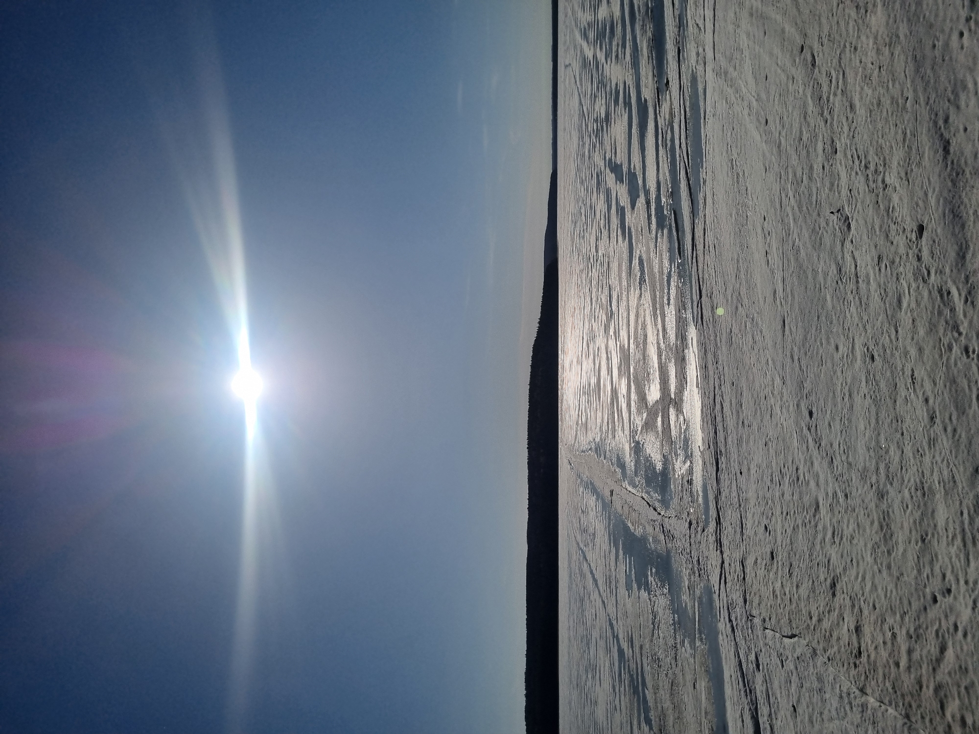 This horizontally oriented image captures a serene, expansive winter landscape, likely a frozen lake or river, under a clear, bright sky. The sun, positioned low in the sky on the left, is a prominent subject, emitting a strong, radiant glare and lens flare that streaks across the blue expanse. Its intense light creates a shimmering, reflective path across the textured frozen surface.
A distant, dark line of coniferous forest forms a subtle horizon, silhouetted against the lighter sky, separating the vast water body from the open air. The frozen surface, which occupies the majority of the lower and right portions of the image, displays intricate textures. It appears to be a mix of ice and compacted snow, marked by numerous cracks, irregular ridges, and areas of varied reflectivity. These patterns suggest a dynamic history of freezing and possibly melting, with some sections appearing smoother and more reflective, particularly where the sunlight hits, while others are rougher and darker. Small indentations or details are visible on the ice, hinting at natural formations or previous tracks.
The scene conveys a sense of cold, crisp weather, with the bright sun indicating either early morning or late afternoon. The overall impression is one of vastness and quiet solitude in a natural, wintry environment. The location context of Chaikovsky, Russia, aligns with the depiction of a frozen landscape. There are no visible people, animals, or discernable artificial objects. No text is visible in the media file.