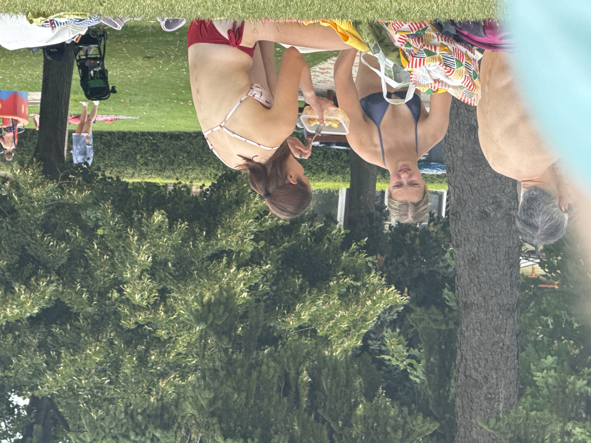The image depicts an outdoor scene in a park in Hainburg an der Donau, Austria, on a grassy area with trees in the background. In the foreground, a man's back is visible, with his head turned to the left. To his right, a young woman in a blue bikini is sitting and looking to the left, with a colorful patterned bag and a yellow towel next to her. Further to the right, another young woman in a white and red patterned bikini is eating from a clear plastic container with a fork, holding it in her right hand. Her hair is tied back in a ponytail. In the middle ground, a woman in a blue shirt and white shorts stands near a tree, and a child is visible in a red and yellow play structure. A black stroller is parked on the grass. The overall setting suggests a recreational area on a sunny day.