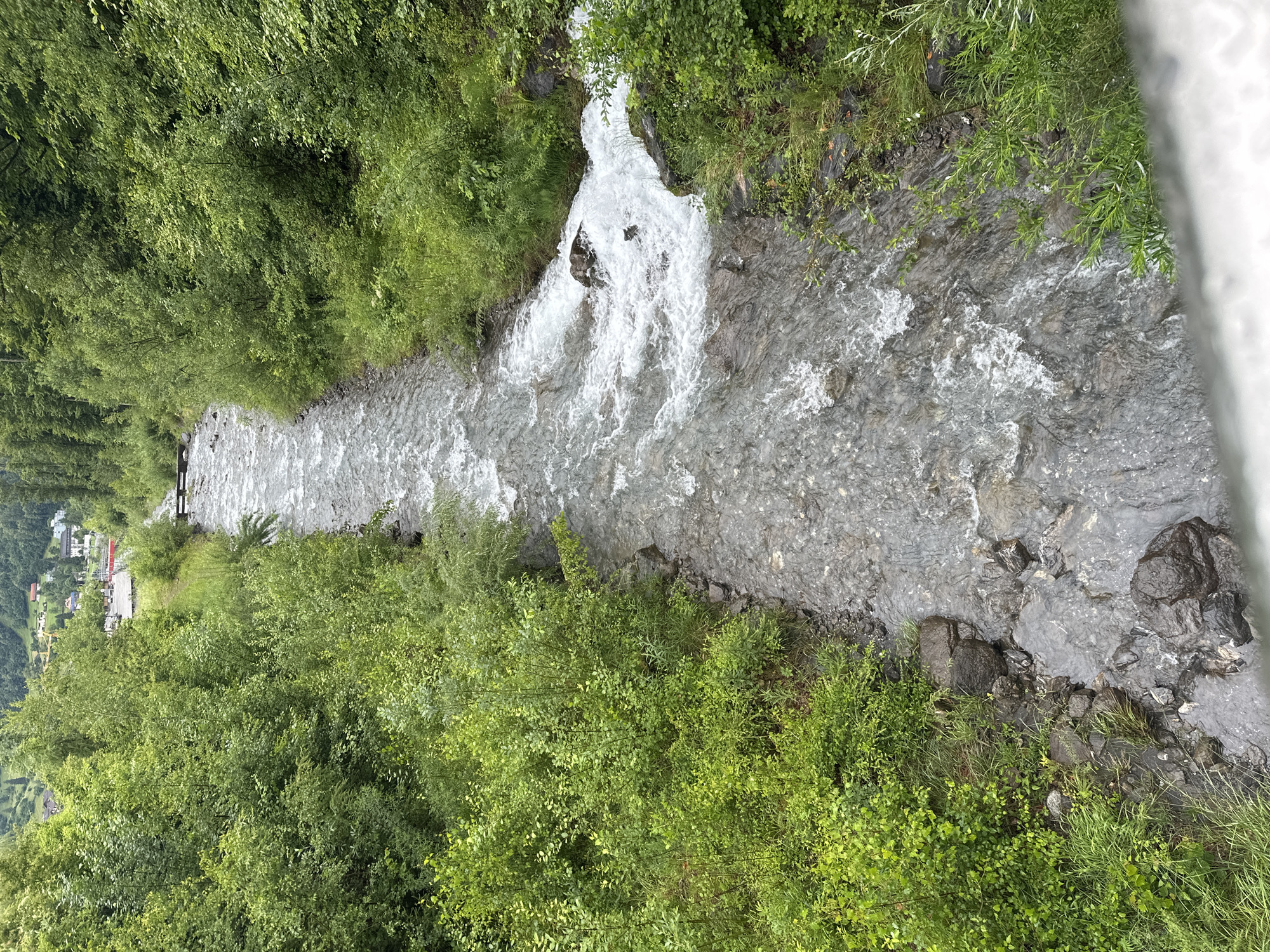 This is an aerial, medium-shot image of a fast-flowing river cutting through a densely wooded valley in Hüsen, Switzerland. The river, filled with rocky debris, is a turbulent grey-blue, with white foam churning over submerged stones. Lush green trees and bushes line both banks, with some taller trees forming a dense canopy further up the slopes. In the distance, a small settlement is visible, nestled among the hills and trees, with a red building and what appears to be a small wooden bridge spanning the river. The overall atmosphere is one of natural, untamed beauty, with the river dominating the landscape.