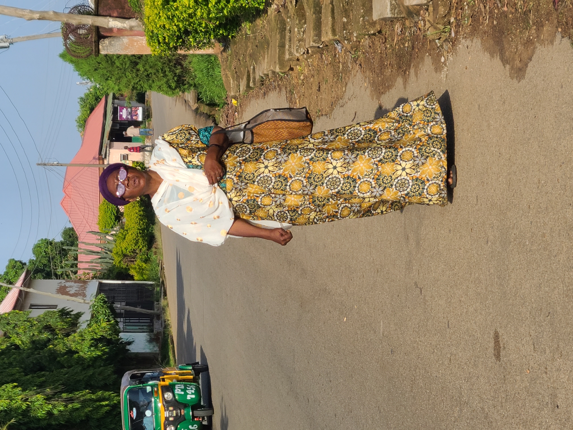 In Jos, Nigeria, a woman is walking on a paved road. She is wearing a purple headwrap, glasses, and a long, yellow and white floral dress. A white shawl is draped over her shoulders. She is carrying a woven bag. To her left, a green and yellow tricycle is parked on the side of the road. The area appears to be a residential street with buildings and lush green trees. The sky is clear and blue, indicating a sunny day.