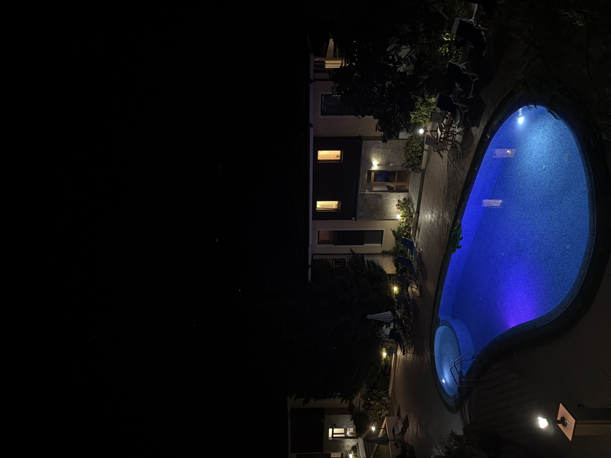 This is a nighttime aerial shot of a swimming pool area with a building in the background. The pool is oval-shaped and illuminated with blue lights, with a smaller circular section. There are several lounge chairs and umbrellas around the pool area. The building in the background appears to be a multi-story structure with illuminated windows. The scene is likely a hotel or resort, possibly located in Sozopol, Bulgaria, given the context. There are no people visible in the image.
