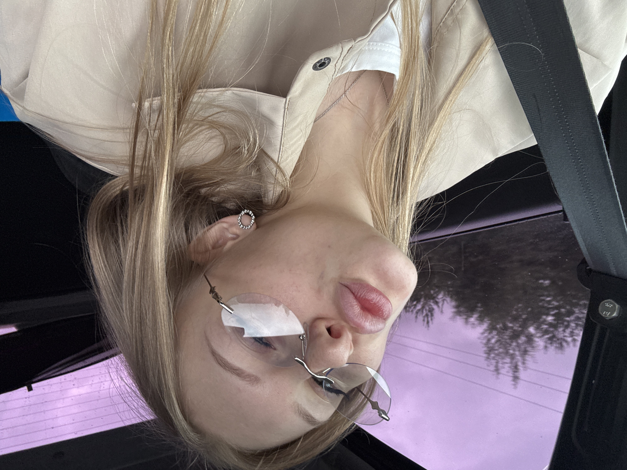 A young woman with blonde hair is inside a car, wearing a light-colored jacket and a white top. She has on unique, rimless glasses and a small, circular earring in her left ear. Her lips are pursed as if she is about to kiss. The background shows a purple-tinted sky with trees visible through the car window. The location is specified as Konratovo, Russia.