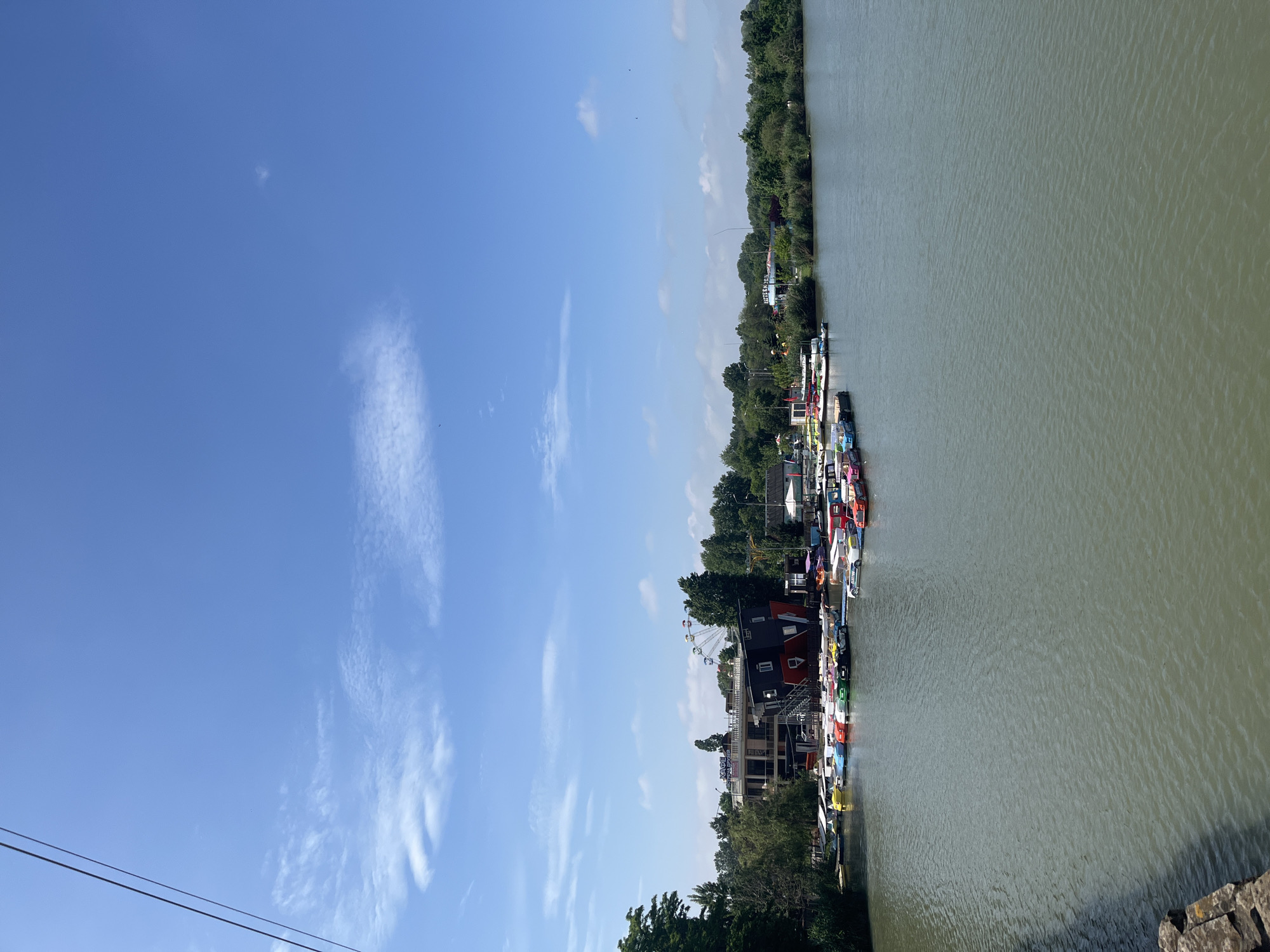 The image shows a waterfront scene in Krasnodar, Russia. The body of water, likely the Kuban River, occupies the right side of the frame. A row of colorful paddle boats is docked along the shoreline. Beyond the boats, a multi-story building with "KAFE SEZONA" signage is visible. A Ferris wheel rises above the building. A line of trees runs along the far bank of the river. The sky is blue with scattered white clouds. In the lower left, power lines are visible.