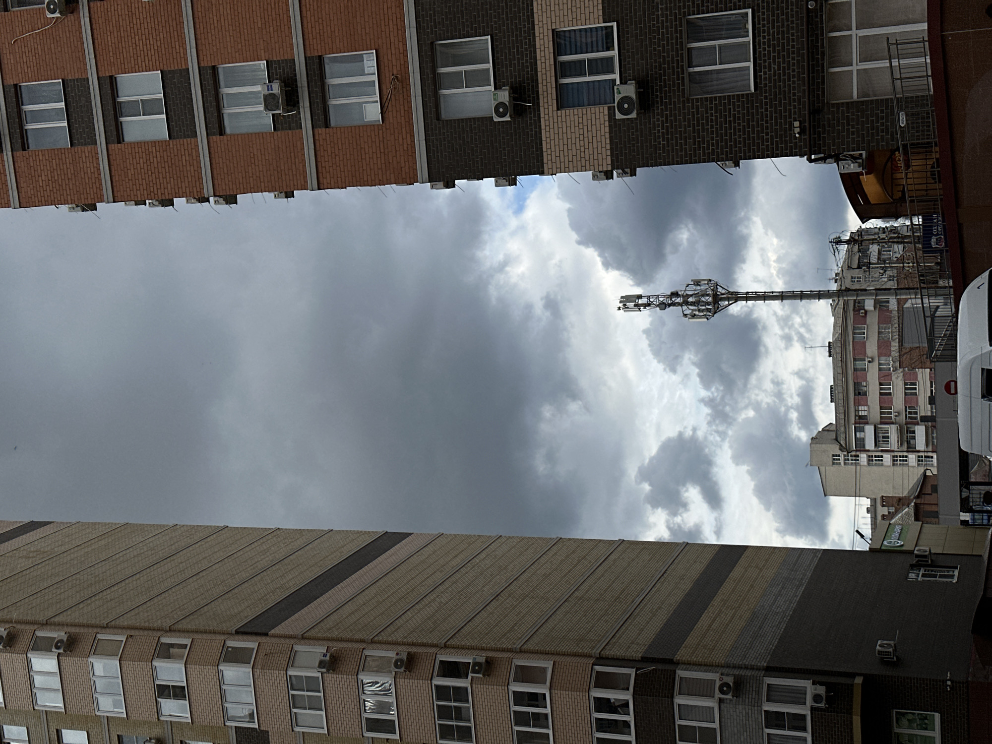 The view is upward from a street in Rostov-on-Don, Russia, framed by the facades of two buildings. Overcast skies fill the space between the buildings. A cell tower is visible against the sky. Other buildings stand in the background. A white van is visible on the right side of the image. Both buildings on the left and right of the scene appear to be residential. Several windows, many equipped with air conditioning units, are visible.