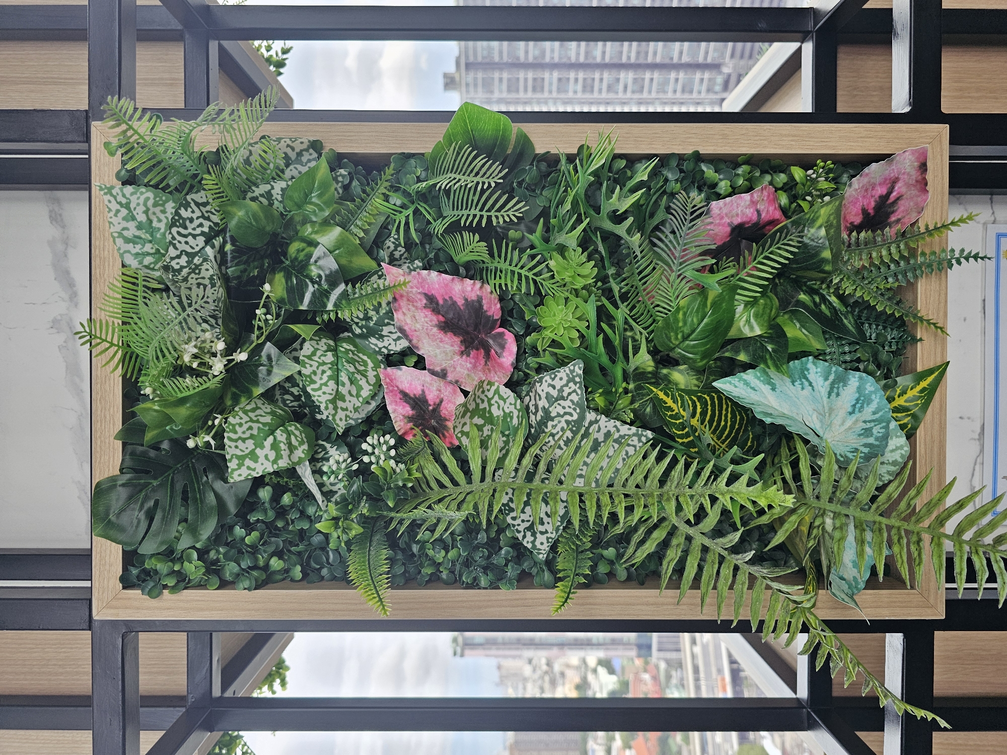 The image shows a vertical artificial plant wall with various green leaves, pink and white patterned leaves, and small white flowers. The plant wall is housed in a light wood-grain frame and is situated outdoors, against a backdrop of a city skyline. In the distance, tall buildings are visible, suggesting an urban environment. The sky is overcast. To the left of the plant wall, a black metal railing and frame are visible, indicating it is likely on a balcony or an outdoor seating area. The overall impression is one of a decorative element in a city setting. The urban landscape appears to be in Bangkok, Thailand, based on the architecture of the buildings and the general cityscape visible. There are no people or animals present in the image.