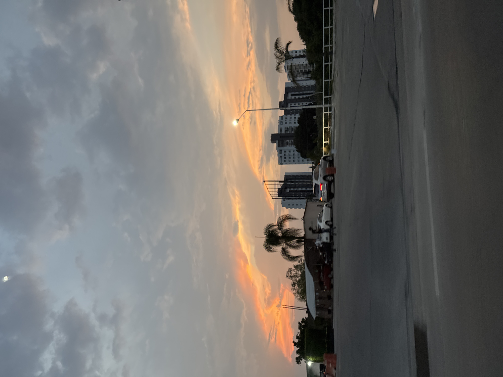 The image shows a street scene at dusk in Puebla, Mexico. The sky is filled with clouds, some of which are illuminated with orange and yellow hues, indicating the setting sun. On the right side of the image, a gray, likely wet, road stretches out, with buildings, including tall apartment blocks, visible in the background. A few cars are parked along the side of the road, and palm trees dot the landscape. A streetlight is illuminated, and there is a suggestion of other urban elements such as fences. The image is taken from a slightly elevated position, facing towards the horizon.