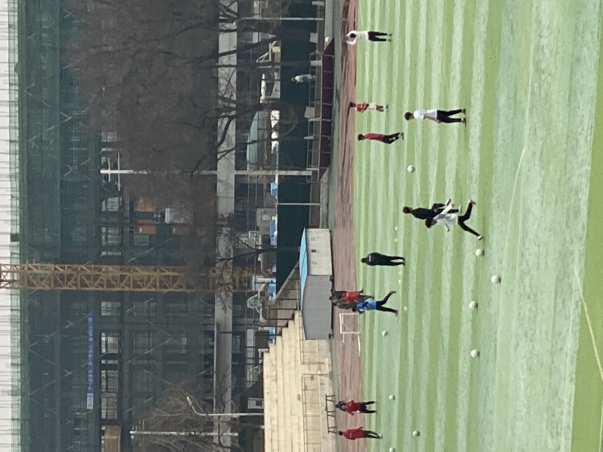 The image shows a soccer field in Zhongtun, China, with several people training or playing. The field is green with white lines, and there are multiple soccer balls scattered around. To the left of the field, there's a stadium with visible seating. Beyond the stadium, there's a construction site with a crane and a building under construction. The weather appears overcast.