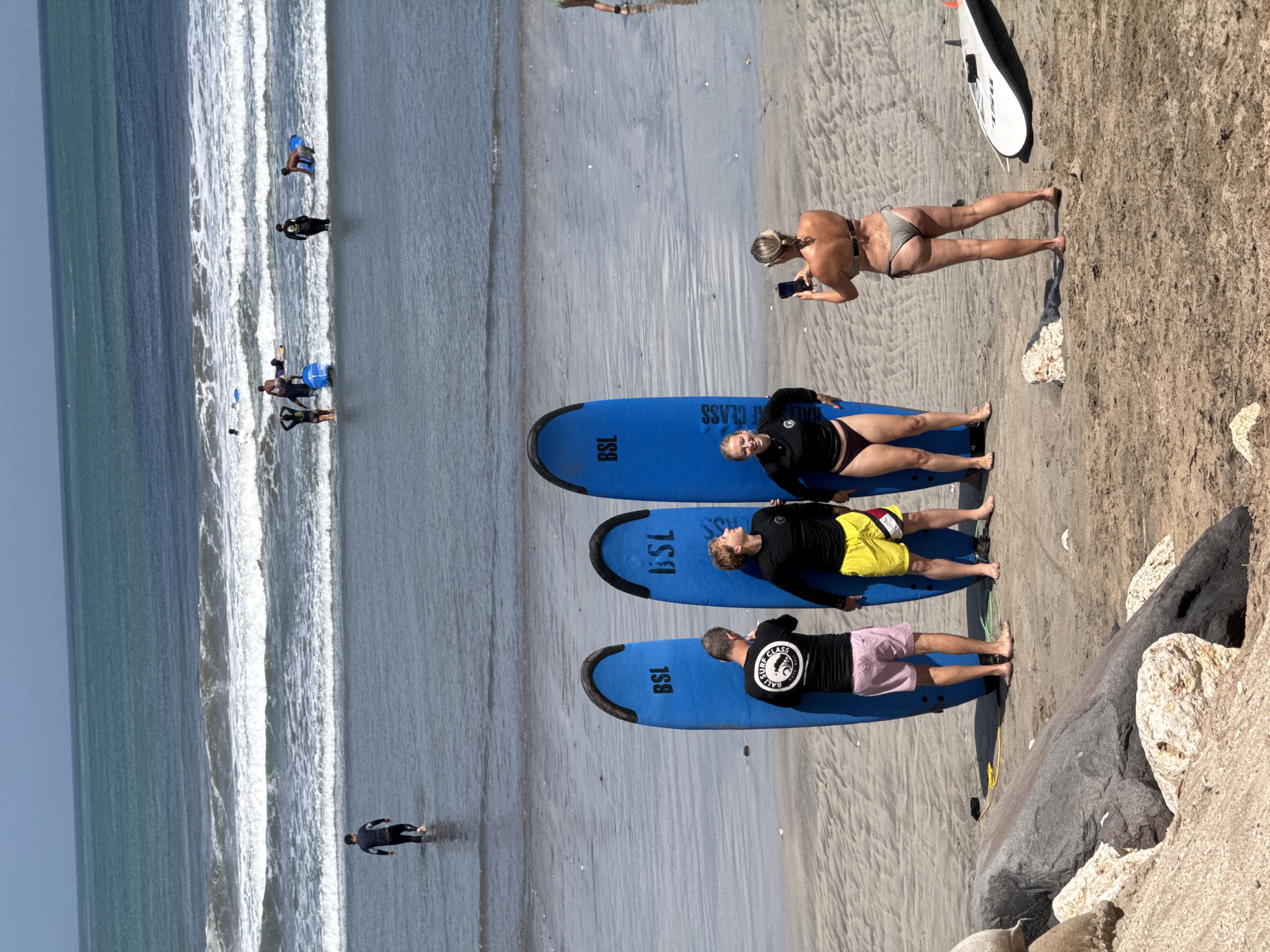 In Kuta, Indonesia, three blue surfboards stand upright on a sandy beach, with the inscription "BSL" visible on two of them. A man in a black long-sleeved shirt and pink shorts stands to the left of the surfboards, facing them. To his right, a man in a black long-sleeved shirt and yellow shorts is looking upwards, and a woman in a black long-sleeved top and burgundy bottoms is smiling at the camera. A woman in a bikini is taking a photo of them with her phone. In the background, waves crash on the shore, and several people are seen surfing or wading in the water, some with surfboards. The sky is a clear blue.