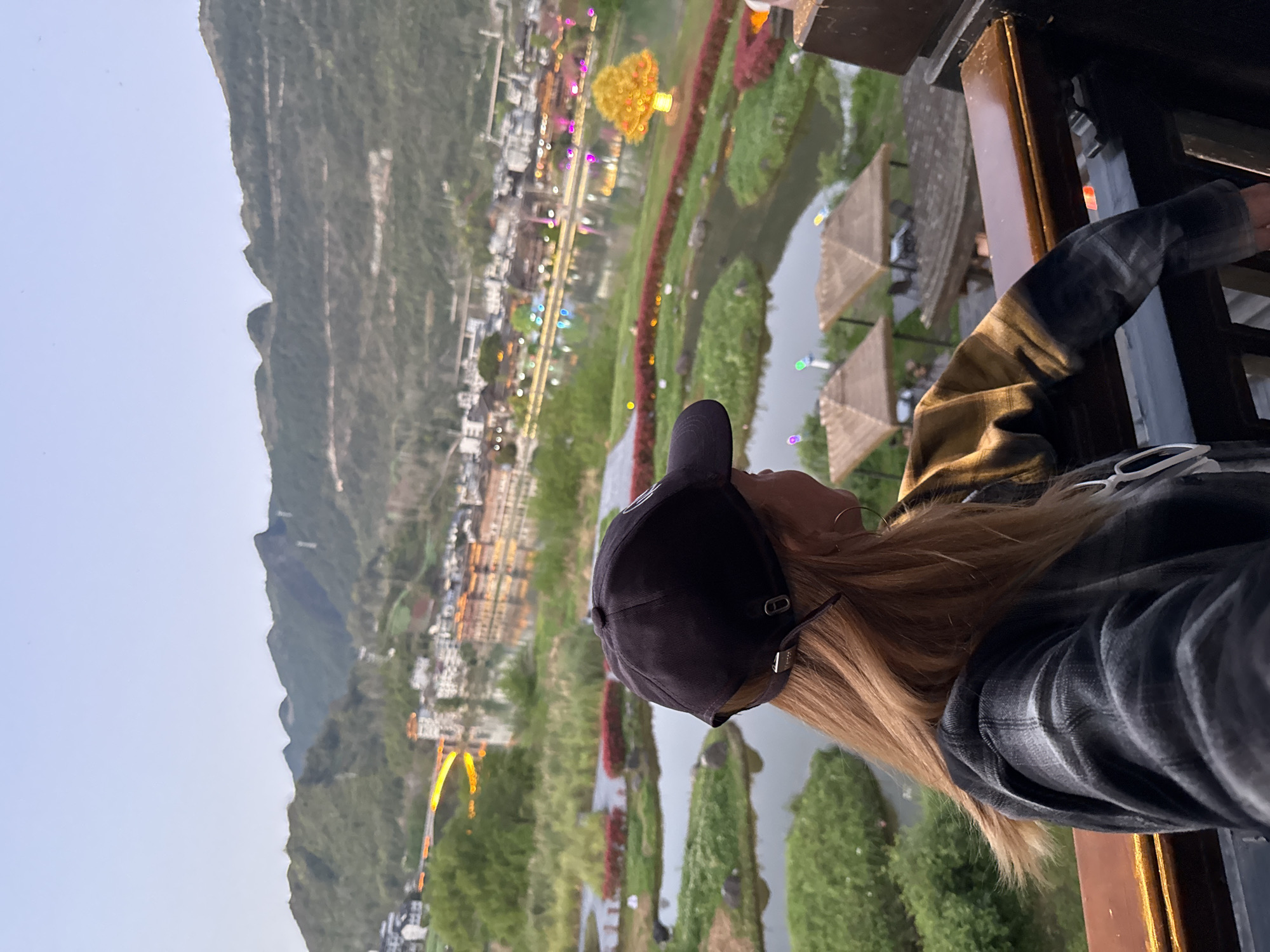 A person wearing a dark baseball cap and a plaid shirt is looking at a scenic view in Zhuoshui, China. The landscape includes a town nestled against a mountain range, a river winding through the town, and decorative structures, including covered walkways and colorful light displays. The person is positioned in front of a window or railing.