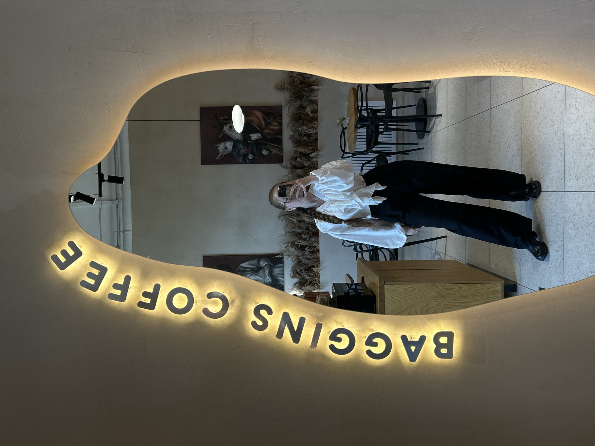 A young woman with a long blonde braid is seen taking a mirror selfie in front of a mirror that reads "BAGGINS COFFEE". The interior of the cafe is visible in the mirror, revealing decorative paintings on the wall and chairs. The scene appears to be set indoors in what is likely a commercial establishment. The exact location within the city of Saint Petersburg, Russia is not discernible from the image alone.