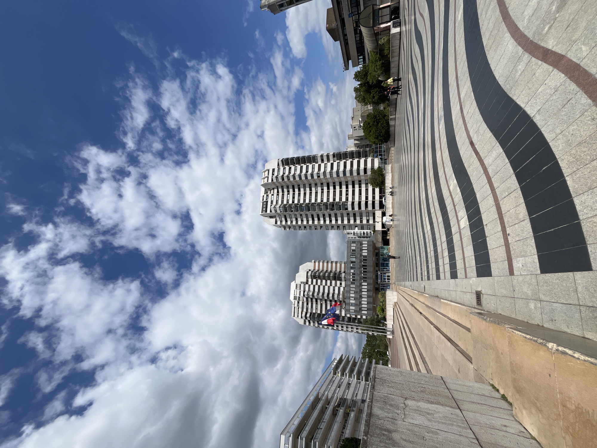 The image captures an outdoor scene with a modern architectural style, likely in Créteil, France. The foreground features a patterned pavement with wavy lines of black, grey, and reddish-brown tiles, leading towards a concrete structure with multiple steps. Beyond this, two tall, white, multi-story buildings with numerous windows stand against a bright blue sky dotted with fluffy white clouds. Flags, including what appears to be the European Union flag and the French flag, are visible near one of the buildings. Trees and other buildings are present in the background, suggesting an urban or semi-urban environment. A few people are seen walking on the pavement and near the buildings. The lighting suggests it is daytime, likely mid-morning or afternoon, with clear, partly cloudy weather. The overall impression is of a clean, contemporary public space. There is no visible text in the image.