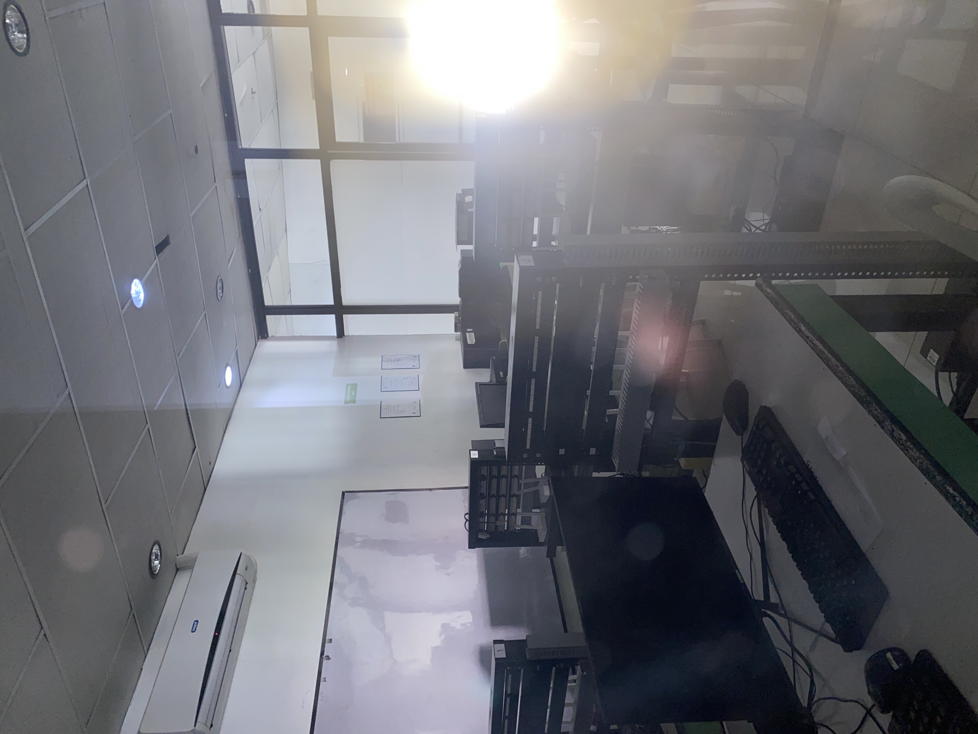 The image displays the interior of a brightly lit computer laboratory or classroom, likely in an educational or professional setting. The room is equipped with multiple computer workstations, each featuring a black monitor, keyboard, and mouse, arranged on light-colored desks. Dark, multi-tiered racks or shelving units separate the workstations.
The setting includes a suspended ceiling with recessed lights, several of which are illuminated. A white split-type air conditioning unit is mounted on one wall, below which a large whiteboard or projection screen is visible. Further along the wall, several framed documents or certificates are displayed.
A prominent feature is a large window or glass partition that allows strong natural light into the room, creating significant lens flare and overexposure in the upper portion of the image. This bright light suggests it is daytime. No people are present, indicating a moment of inactivity, though the setup is clearly for computer-based work or learning. Various cables are visible on and around the desks, connecting the equipment. Faint labels, possibly "1" and "C-1," can be seen on some equipment. The overall impression is a functional, organized, and well-lit environment. The presence of the split-type air conditioner is a common feature in the tropical climate of Davao City, Philippines.