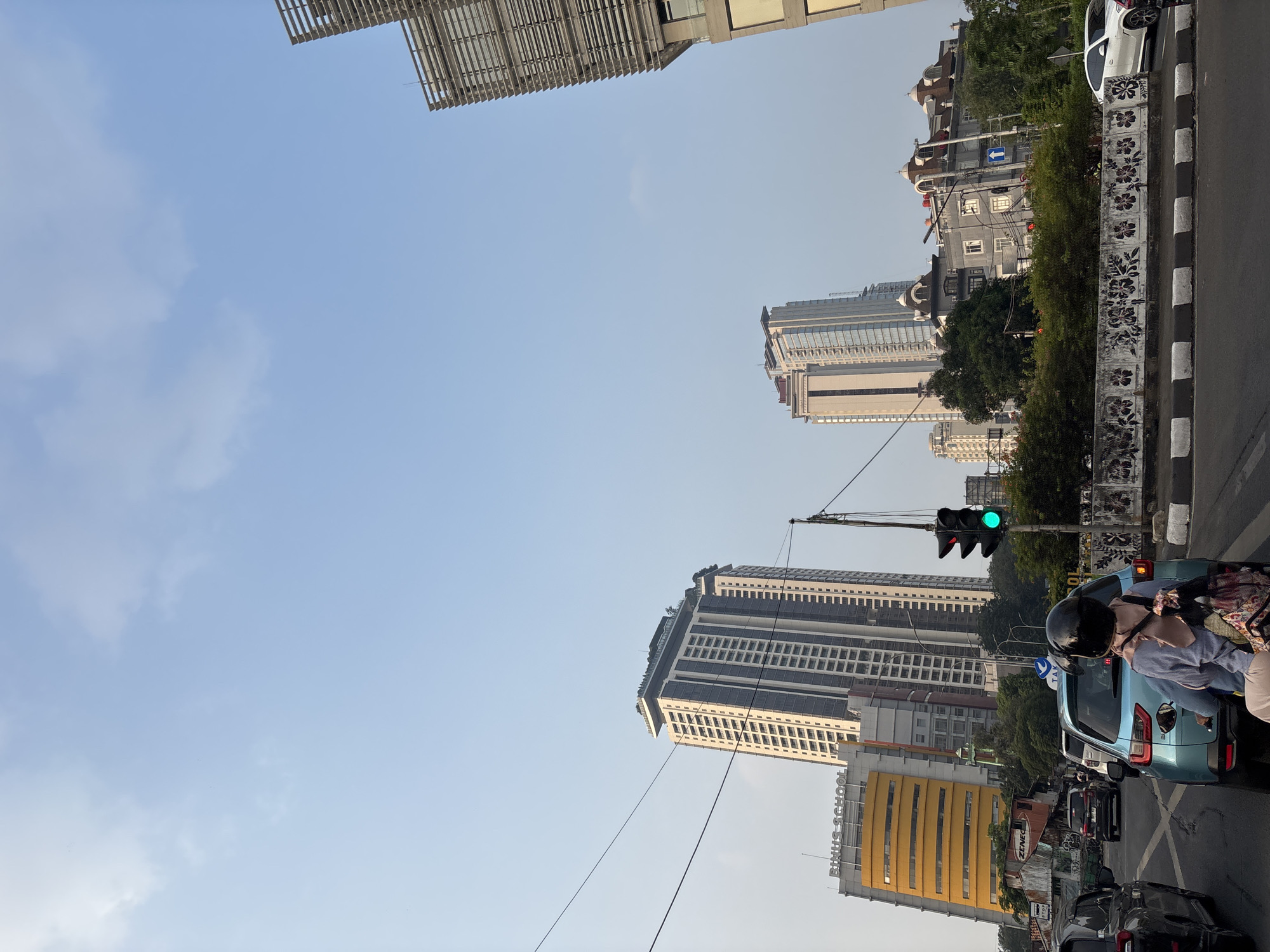 In South Jakarta, Indonesia, on a clear day with a pale blue sky, traffic is at a standstill. A blue car with a rider wearing a light blue top, hijab, and helmet is in the foreground. The rider is facing away from the camera, and their backpack is visible. A traffic light displays a green signal. In the background, the Binus School, a yellow, multi-story building, is visible, as are several tall skyscrapers and other buildings. The environment is urban, with power lines crisscrossing the sky and trees lining the street.