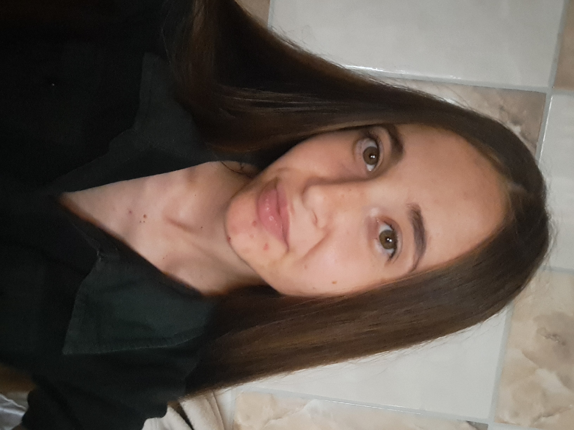 A young woman with long, dark brown hair is pictured in a close-up selfie. She has light skin with a few blemishes and freckles, brown eyes, and light pink lips. She is wearing a dark collared shirt. The background appears to be a tiled wall with light-colored square tiles. The image is taken indoors, likely in a residential setting, and does not provide any specific visual cues to identify its location within Vypasne, Ukraine.